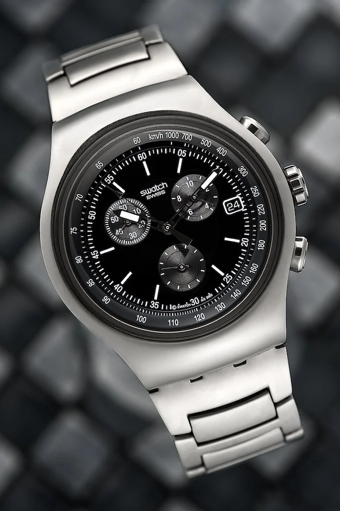 A silver wristwatch with a black dial, multiple sub-dials, and a date window, resting on a dark, textured surface.