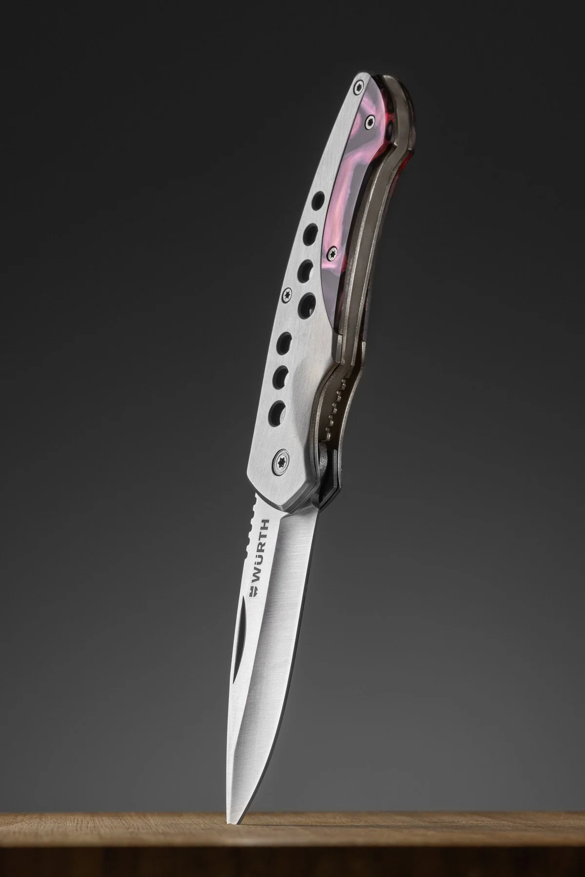 A folding pocket knife with a partially serrated blade, displaying the WÜSTH logo, standing upright on a wooden surface against a dark background.