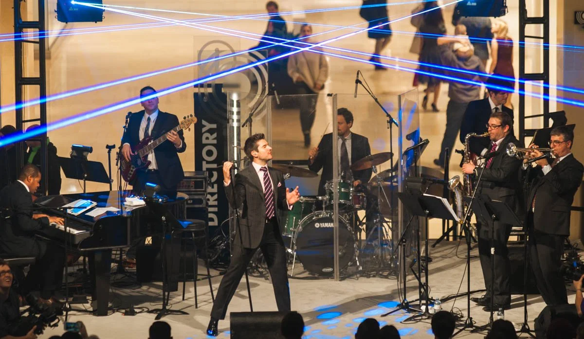 A musical band performing on stage with a lead singer, pianist, drummer, bassist, and saxophonists, in front of an audience with blue laser lights overhead.
