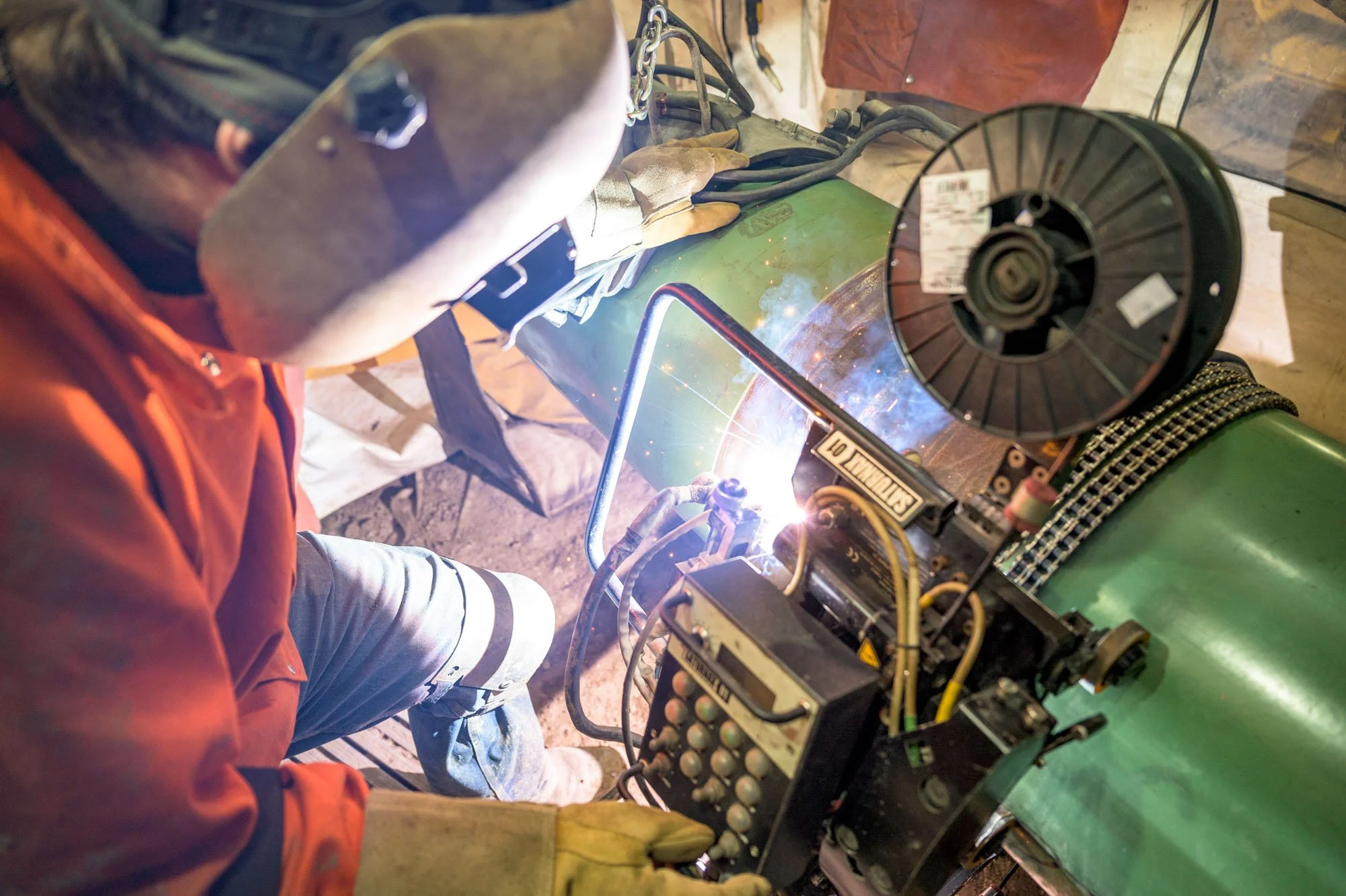 A person welding metal using a welding machine, wearing protective gloves and mask, with bright sparks and light from the welding process visible.