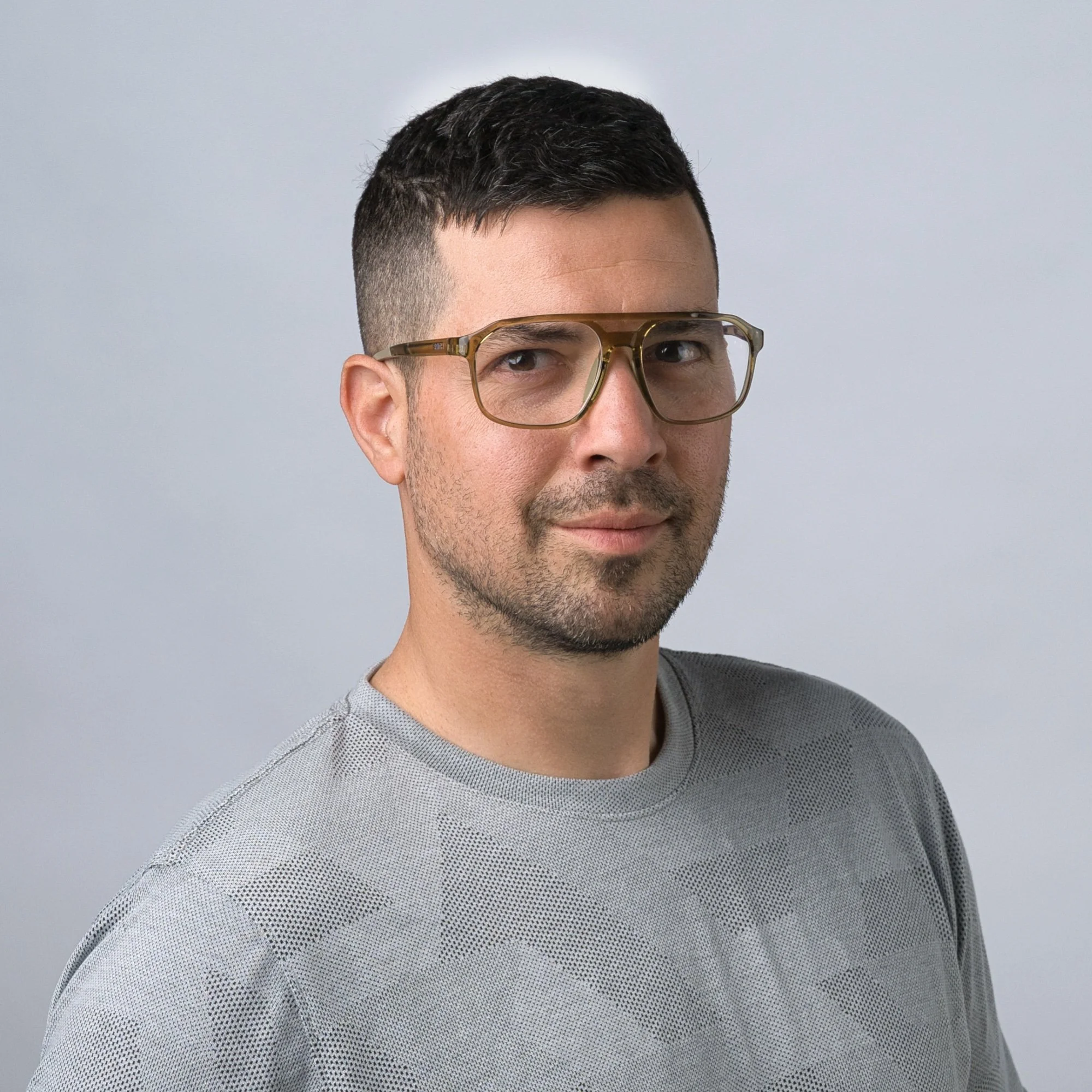 A man with short dark hair, glasses, and a trimmed beard wears a gray patterned shirt against a light gray background.