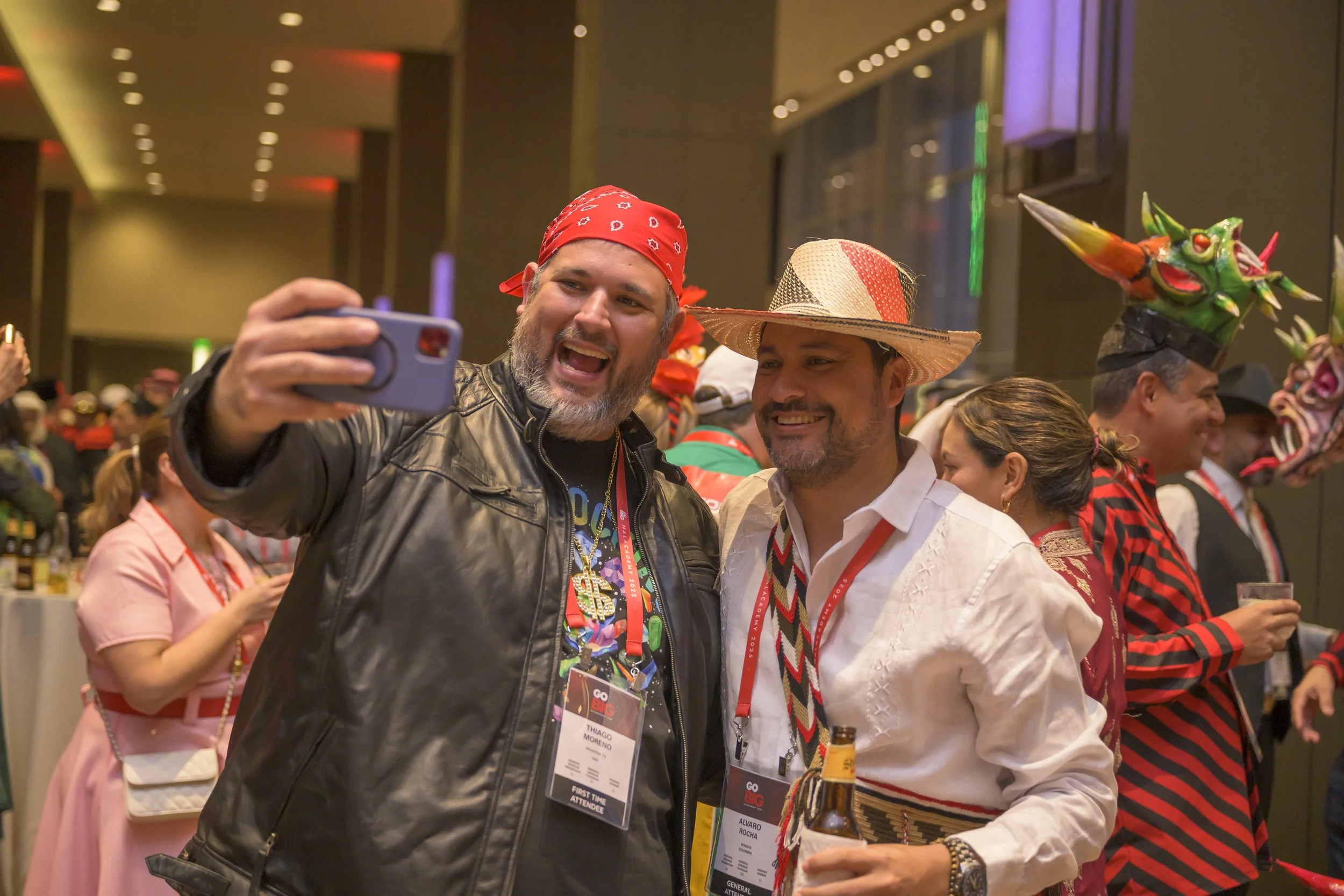 Two men taking a selfie at a festive event, surrounded by people in costumes and colorful decorations.