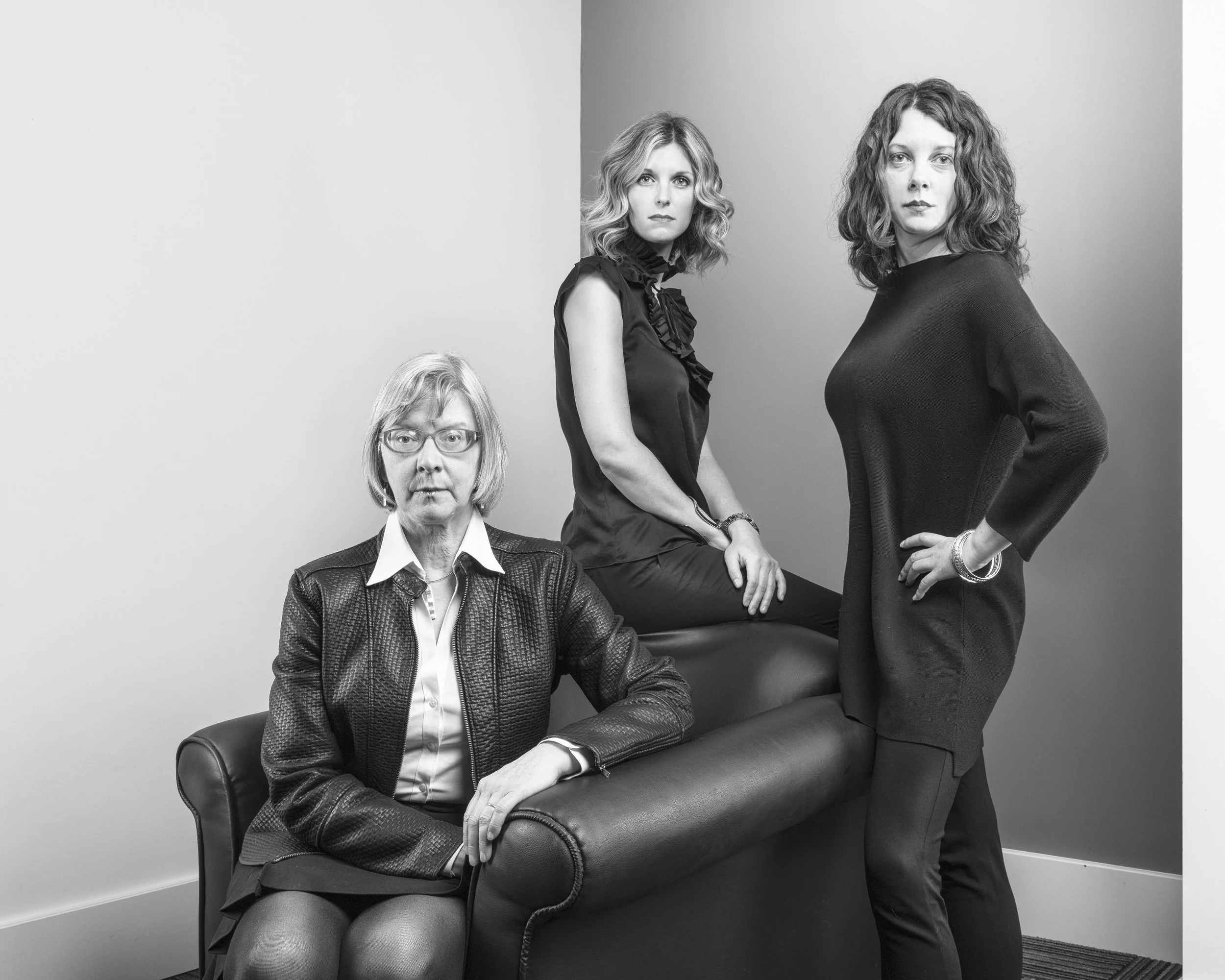 Black and white photo of three women in an office setting. One woman is sitting on a chair with her hand on her knee, wearing glasses and a leather jacket. The other two women are standing, one sitting on the armrest of the chair, both with curly hai