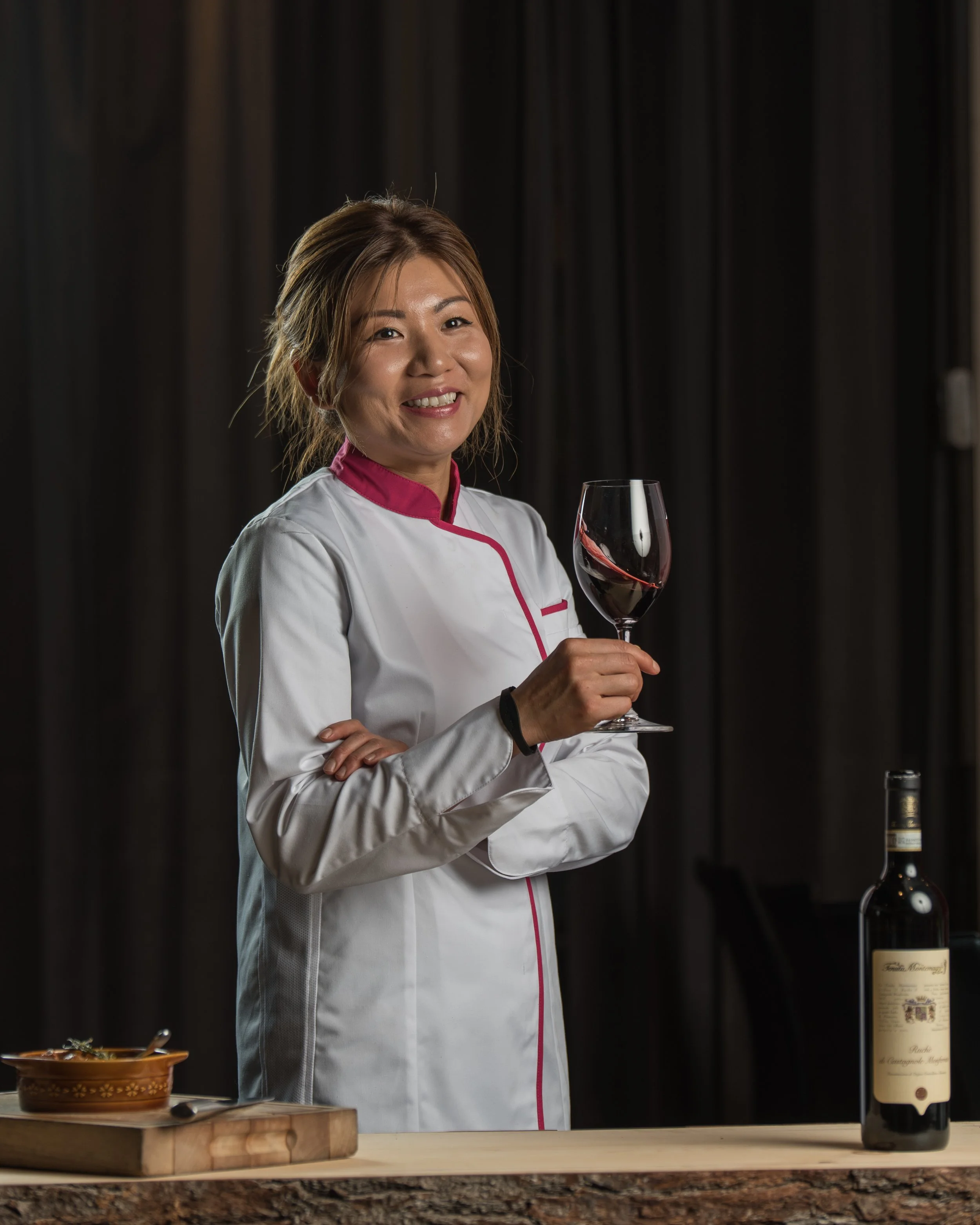 A woman in a chef's coat holding a glass of red wine, standing behind a wooden table with a bottle of wine and a loaf of bread.