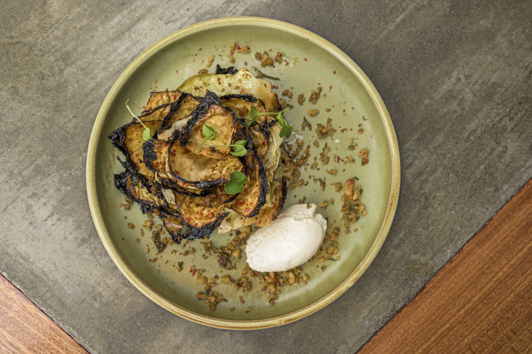 A plated dish with grilled vegetables topped with microgreens, a dollop of white sauce or cream, and sprinkled crushed nuts or crumbs on a green ceramic plate.