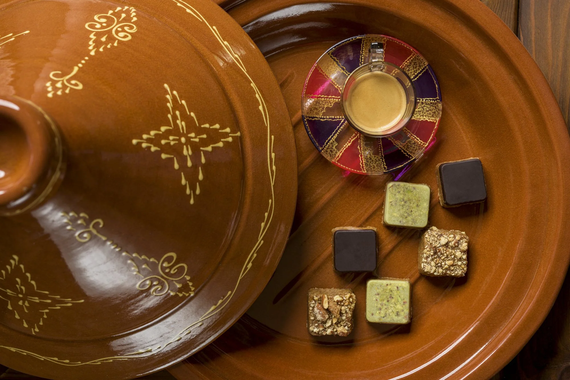 A top-down view of a round wooden tray with a small glass cup filled with a light brown drink, placed on a colorful saucer with gold trim. Surrounding the cup are six small square chocolates, some with toppings, on the tray.