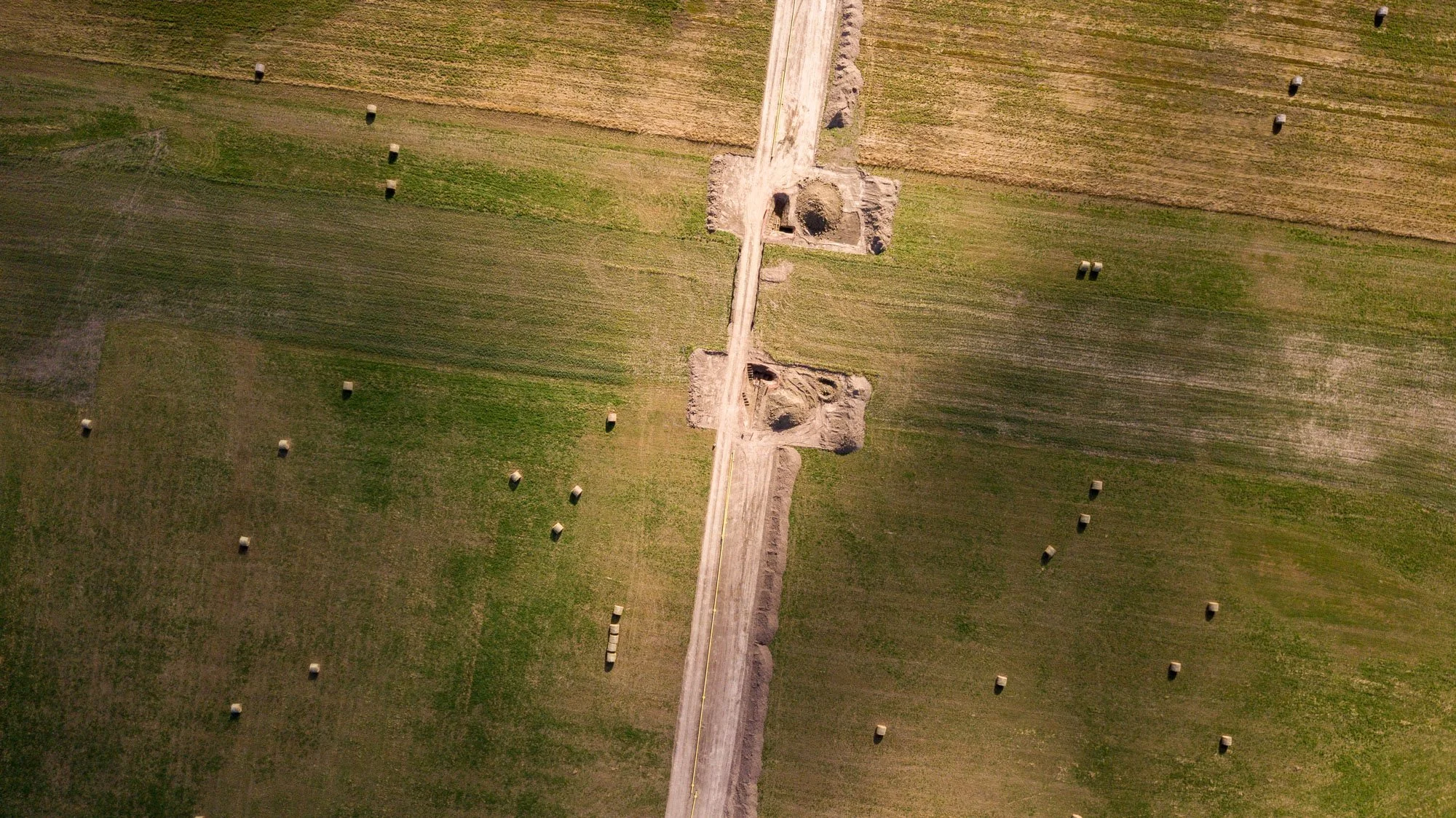 An aerial view of farmland with large rectangular hay bales scattered across green fields. There is a dirt road running through the middle with construction equipment and partially dug holes.