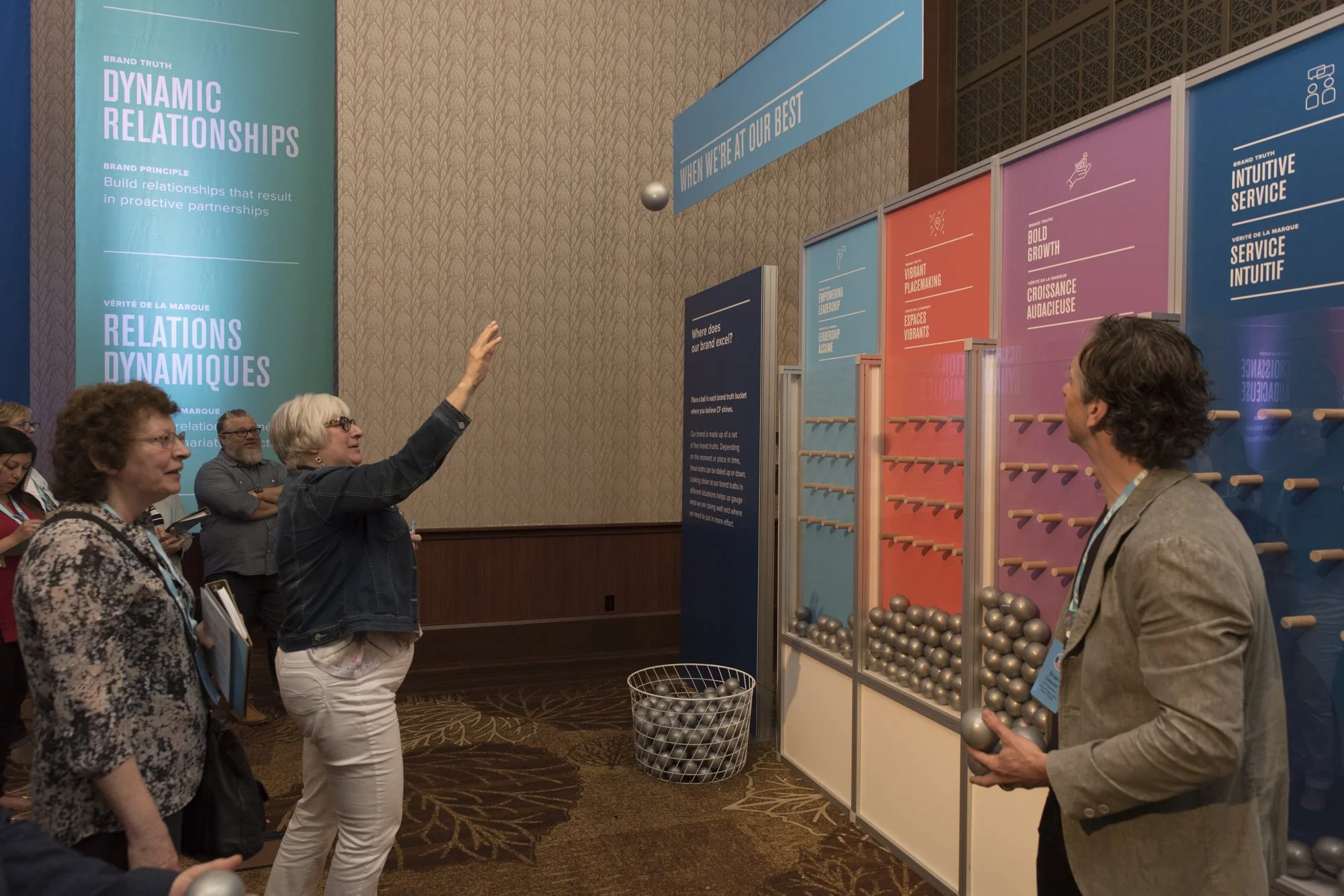 A group of people engaging in a game involving metal balls, with some waiting in line and others tossing balls, in a brightly colored exhibition area with large banners and informational displays about relationships and service.