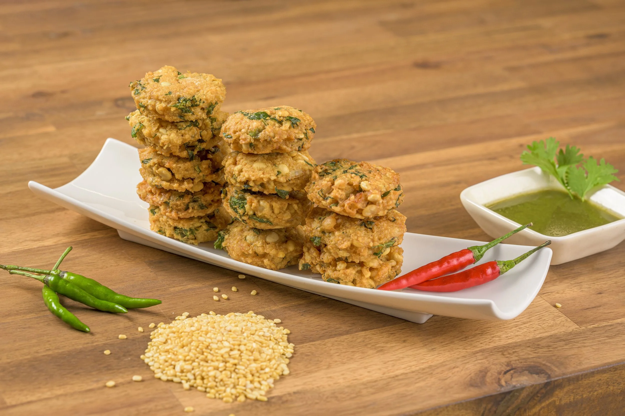 Fried vegetable cutlets stacked on a white rectangular plate with green and red chili peppers, a small bowl of green sauce garnished with coriander, and some sesame seeds on a wooden surface.