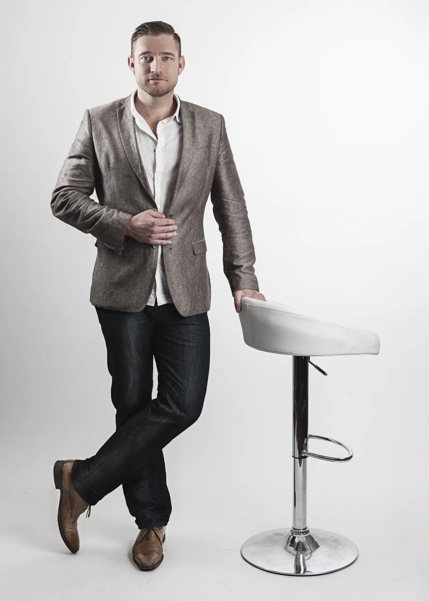A man in a gray blazer, white shirt, and dark jeans standing next to a white bar stool against a plain white background.