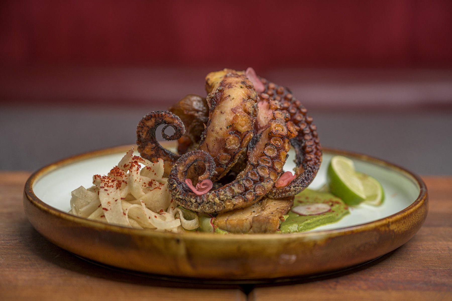 Grilled octopus tentacle with side of pickled vegetables and lime wedge on a ceramic plate.