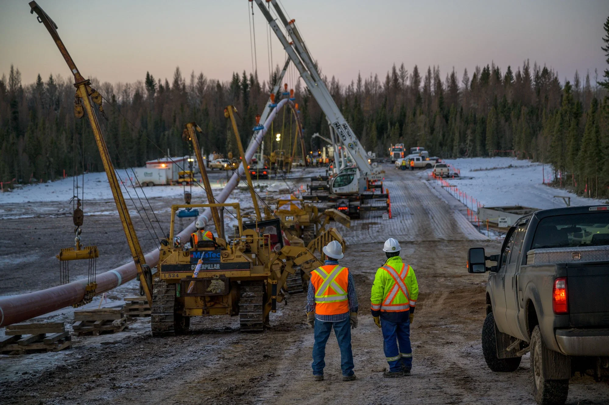 Construction workers wearing safety vests and helmets oversee the installation of large utility poles on a snowy, dirt road, with crane equipment and trucks in the background.