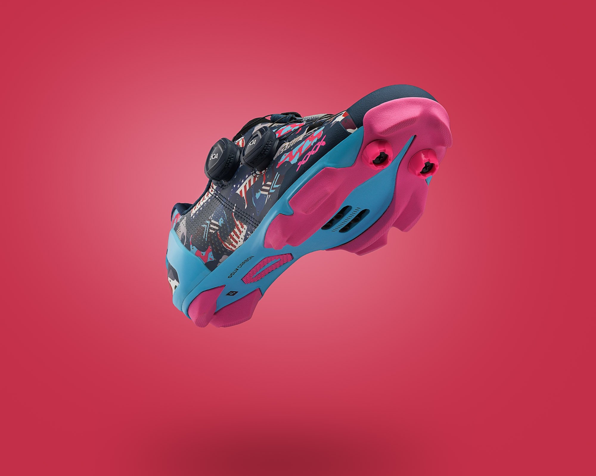A colorful cycling shoe with pink, blue, black, and white details, featuring a vibrant pattern and two black dial closures, suspended against a pink background.