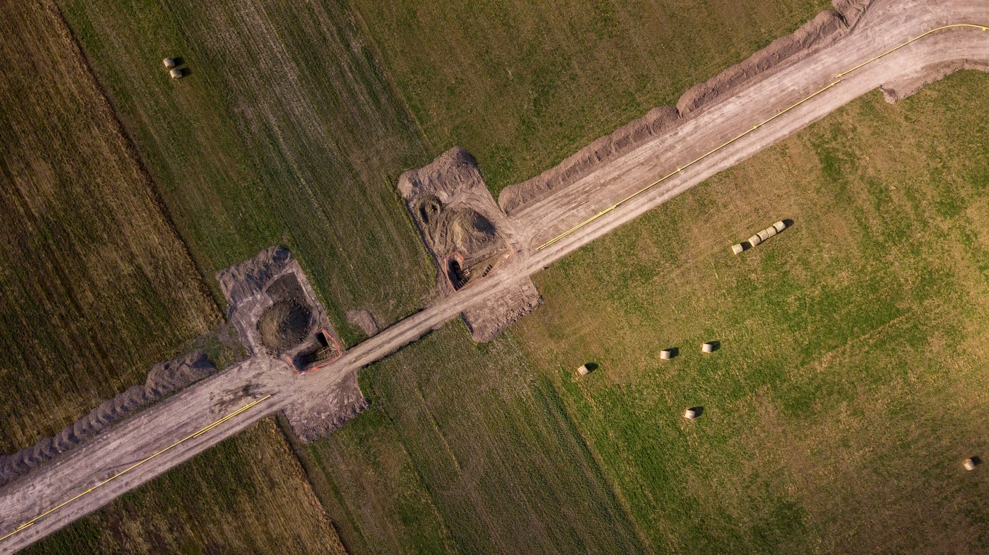 Aerial view of a rural construction site with two excavated areas, a dirt road, and hay bales in a green field.