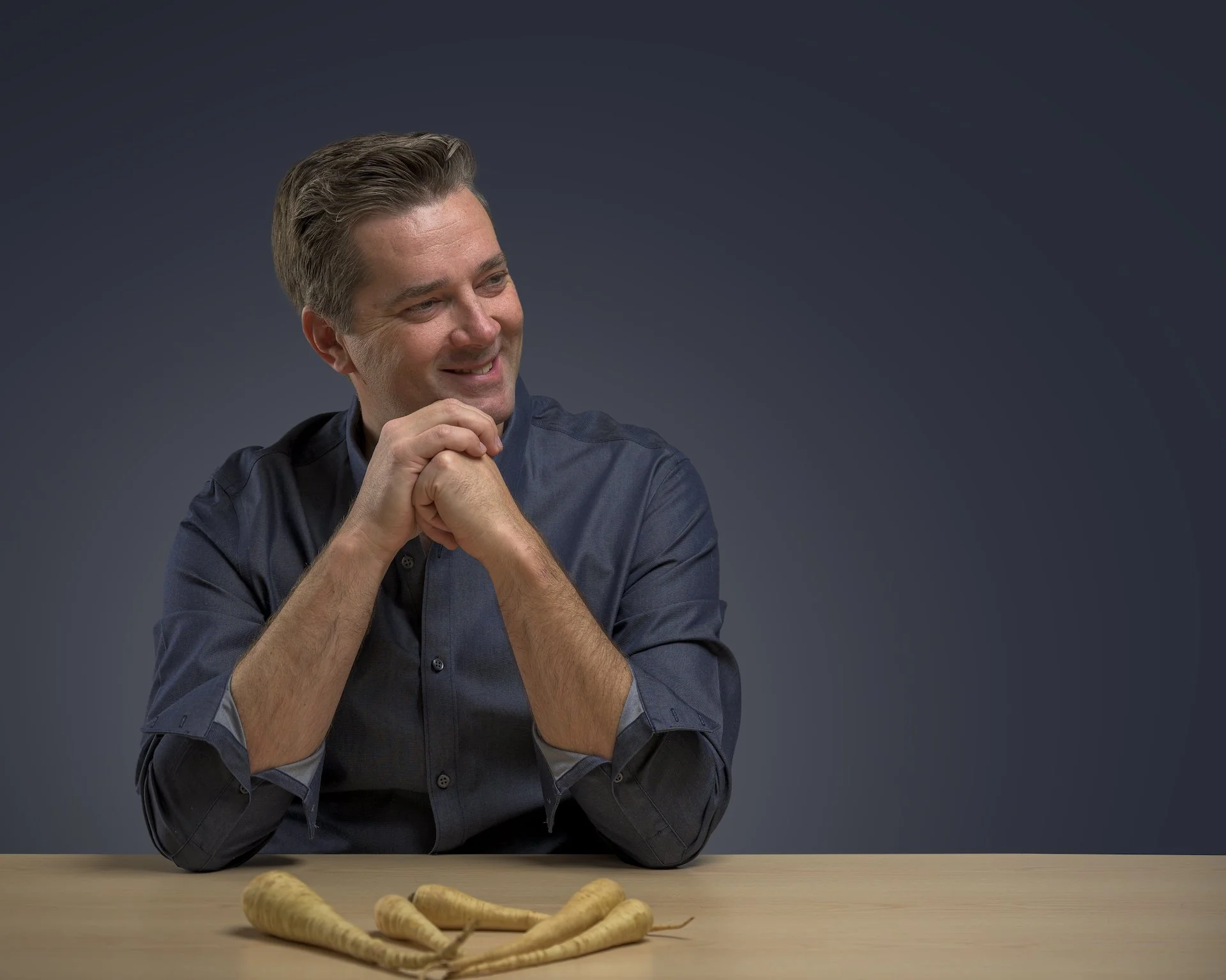 A smiling man with gray hair sitting at a table with several ginseng roots in front of him, against a dark gray background.
