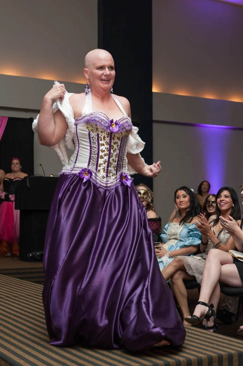 A woman with a shaved head wearing a purple and white cosplay dress, smiling, at an indoor event with an audience.