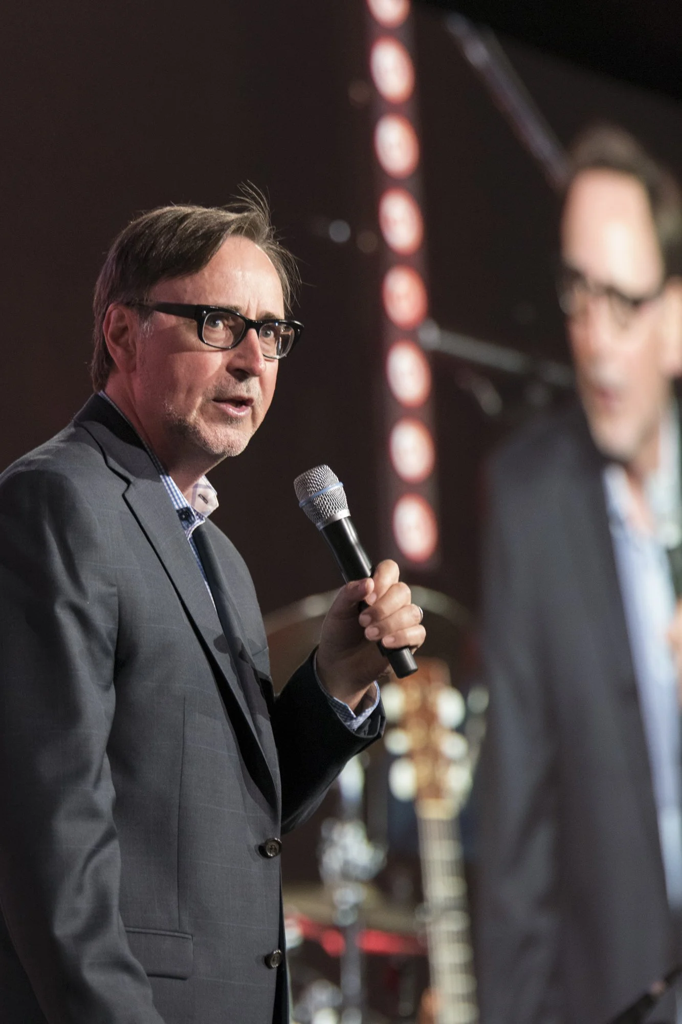 Man in a gray suit and glasses holding a microphone speaking on stage, with a large screen displaying his image and blurred background including musical instruments.