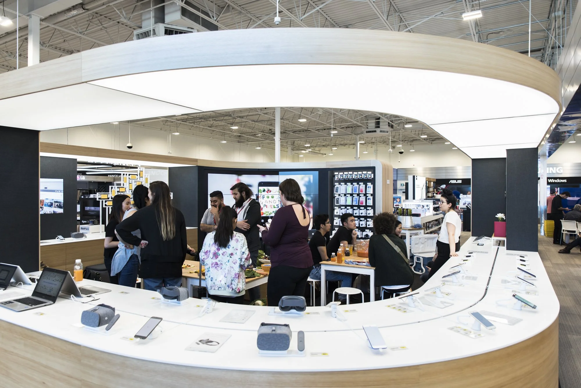 People shopping for electronics inside a retail store, with smartphones and virtual reality headsets displayed on a curved white counter.