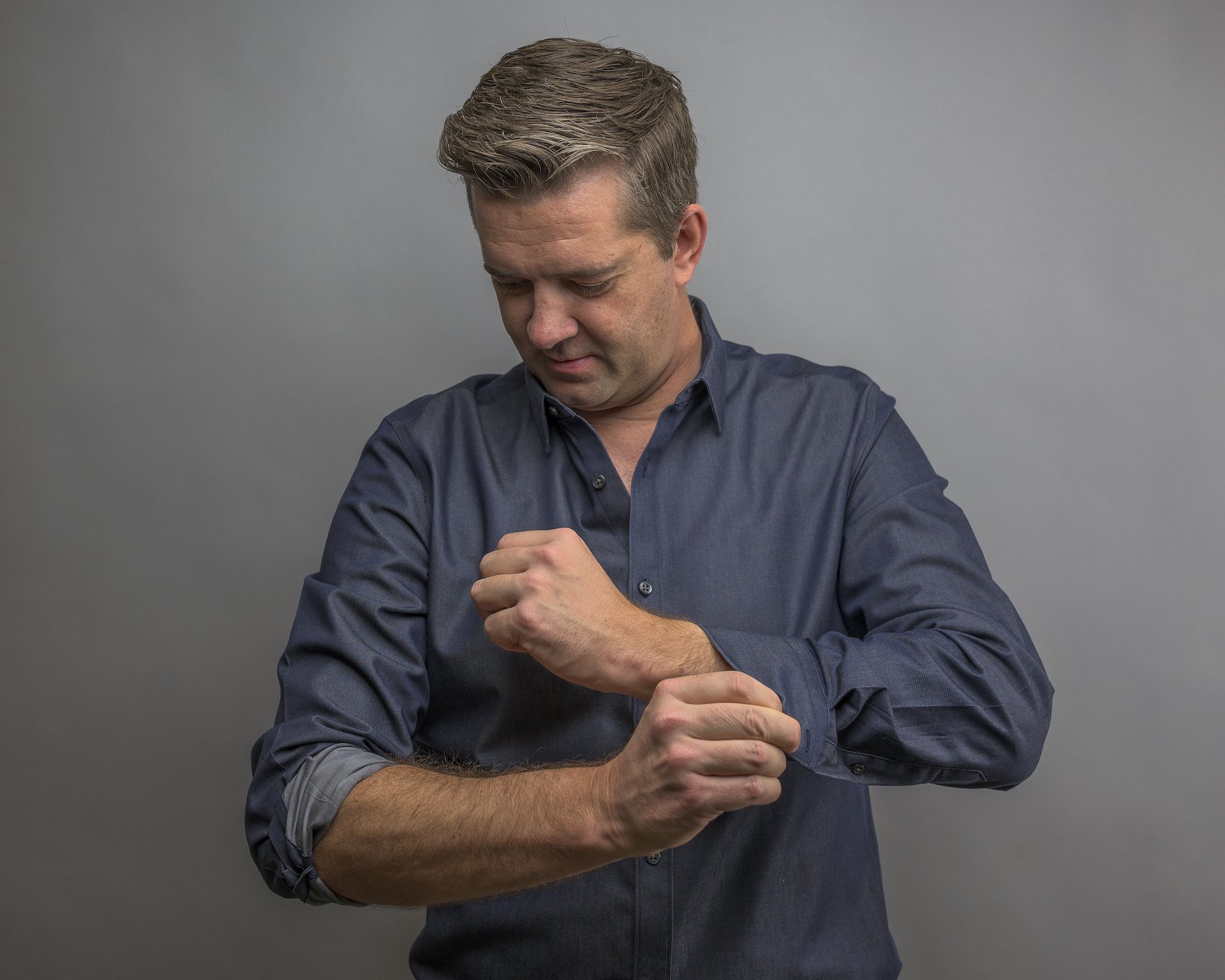 A man in a blue shirt rolling up his sleeve to put on a watch or band.