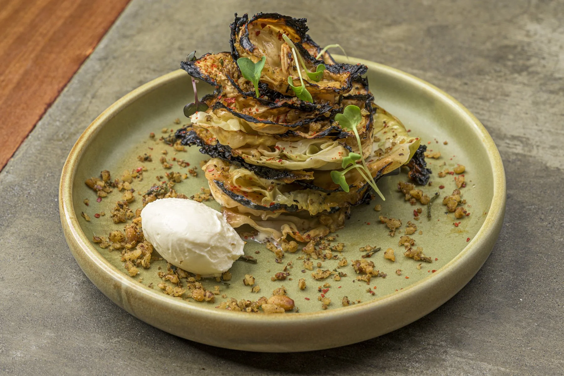 Stack of grilled vegetables garnished with microgreens, served with a side of creamy sauce and sprinkled with crushed nuts or crumbs on a ceramic plate.
