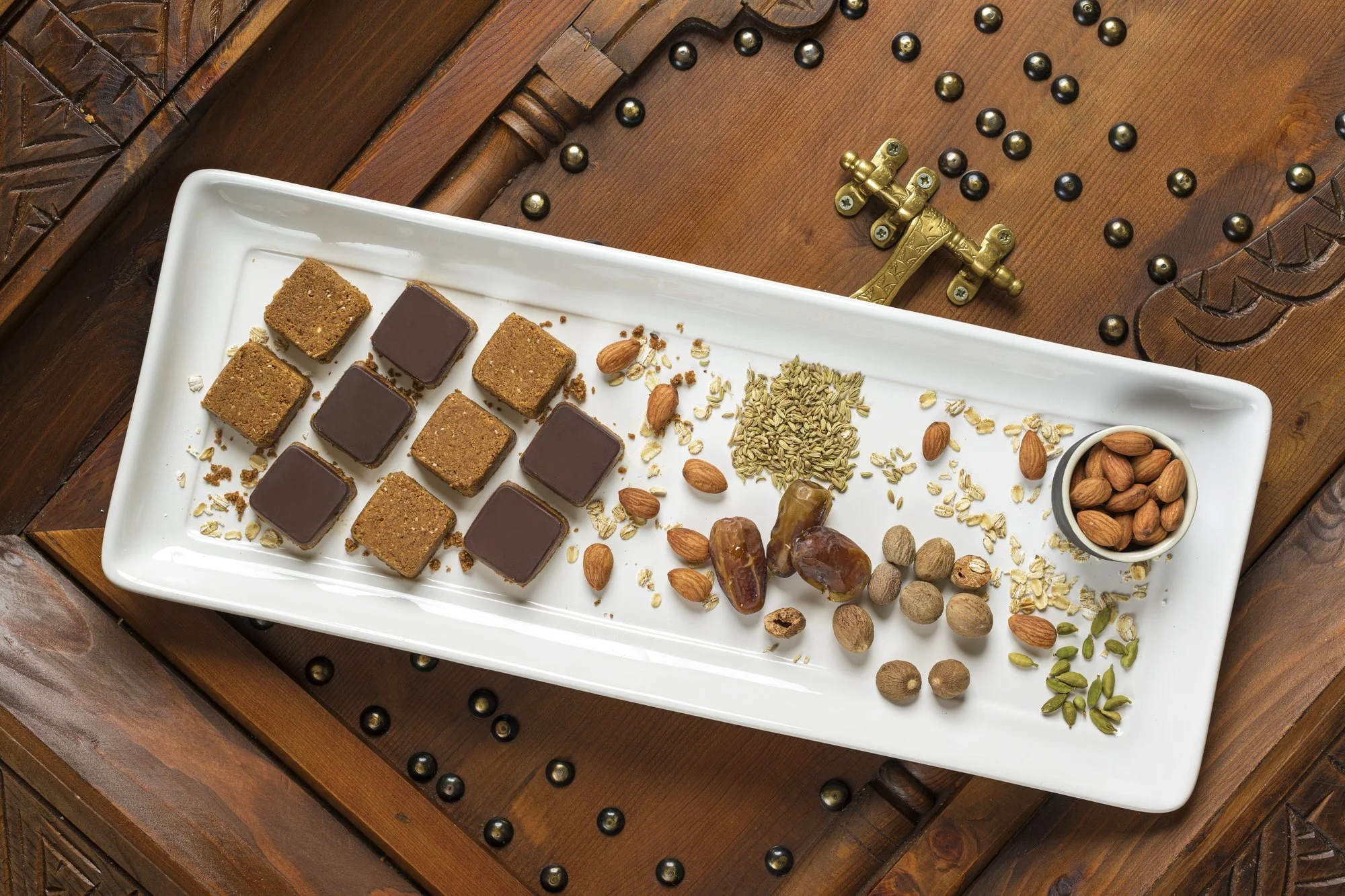 A white rectangular platter containing assorted Indian sweets and nuts, set on a wooden table with decorative brass latch and black beads around it.
