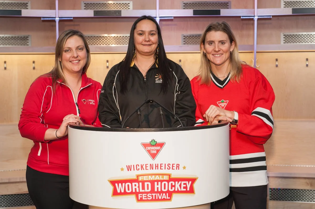 Three women standing behind a podium with a sign that reads 'Canadian Tire Wickenheiser Female World Hockey Festival'.
