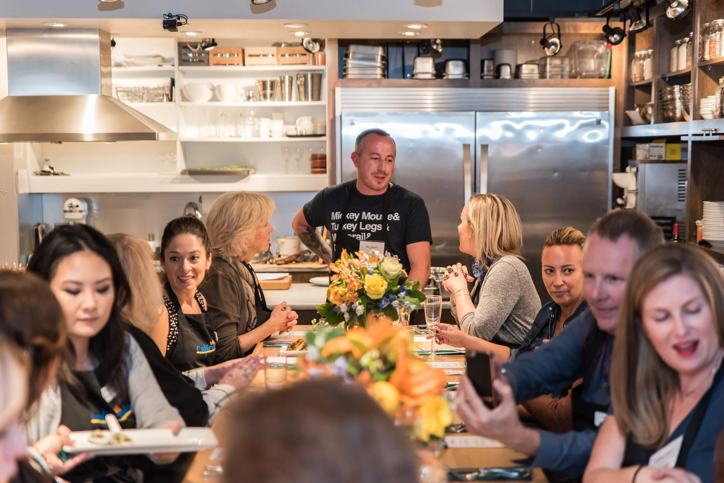 Group of people seated at a table in a kitchen, engaging in conversation during a social event, with a man standing and talking at the center and a floral centerpiece on the table.