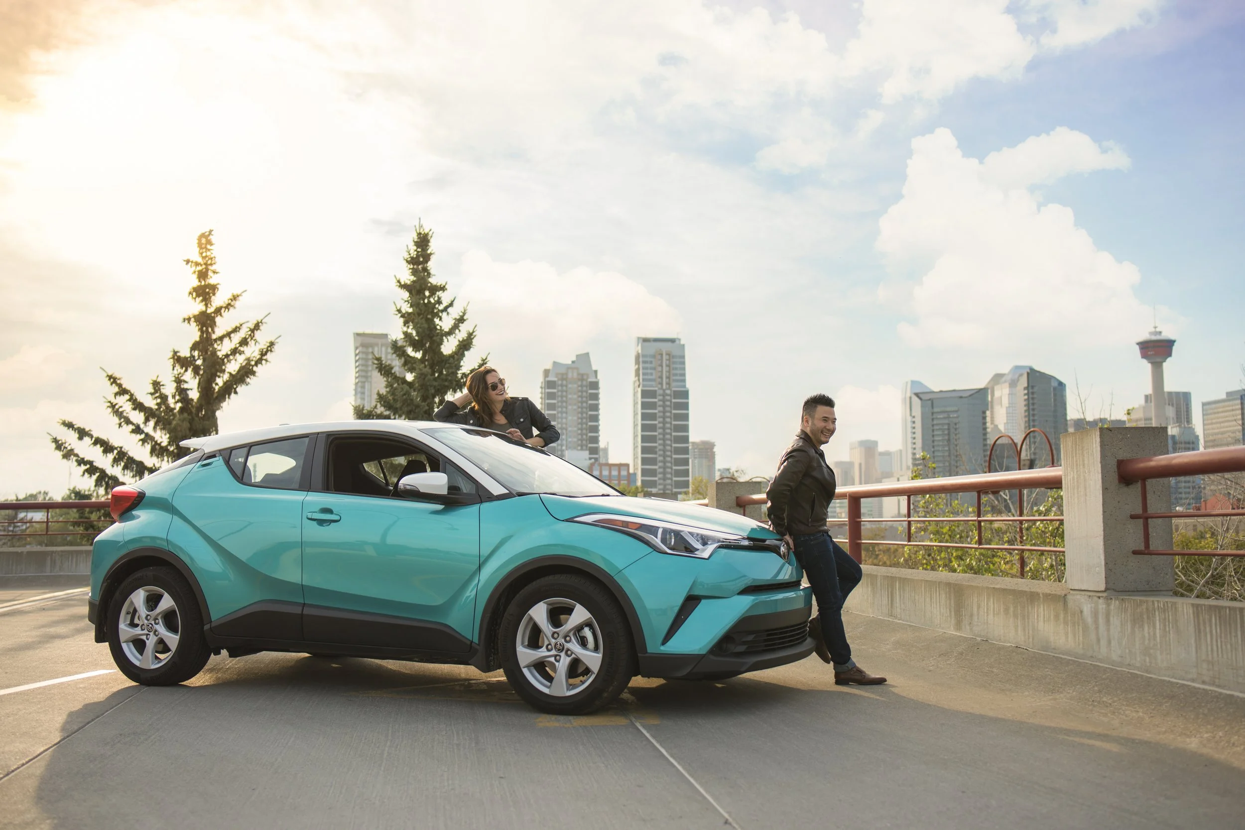 Two people enjoying a moment outside a teal compact car in an urban parking lot. One person is sitting on the car's roof, and the other is leaning against the front of the vehicle. The city skyline is visible in the background under a partly cloudy s