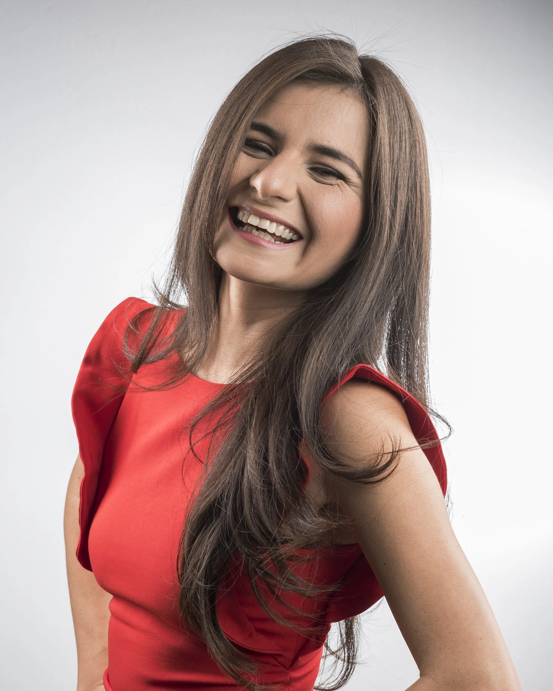 Woman in a red dress smiling and showing her teeth, with long brown hair and a white background.