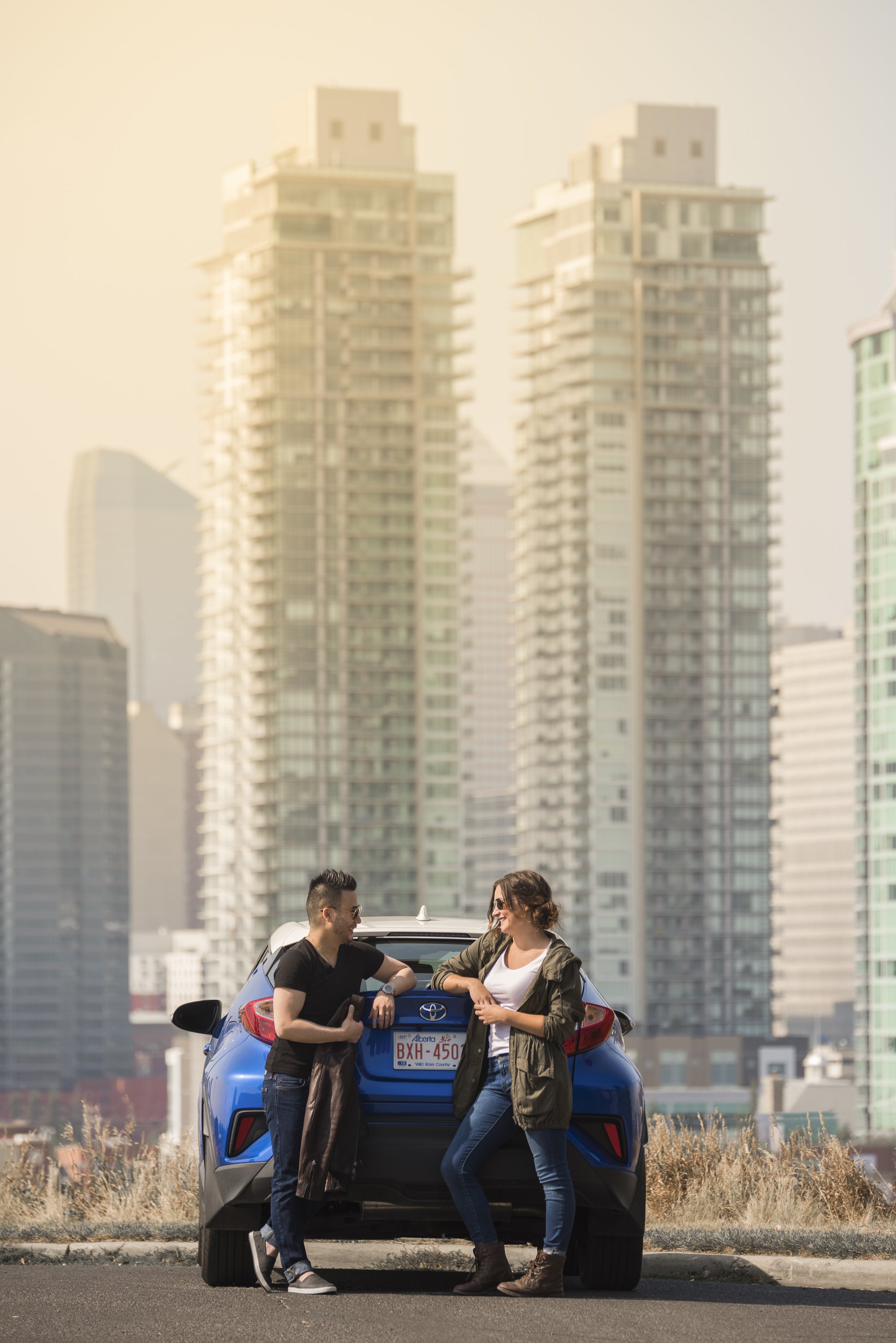 A man and a woman talking and smiling in front of a blue Toyota car with a city skyline of high-rise buildings in the background.