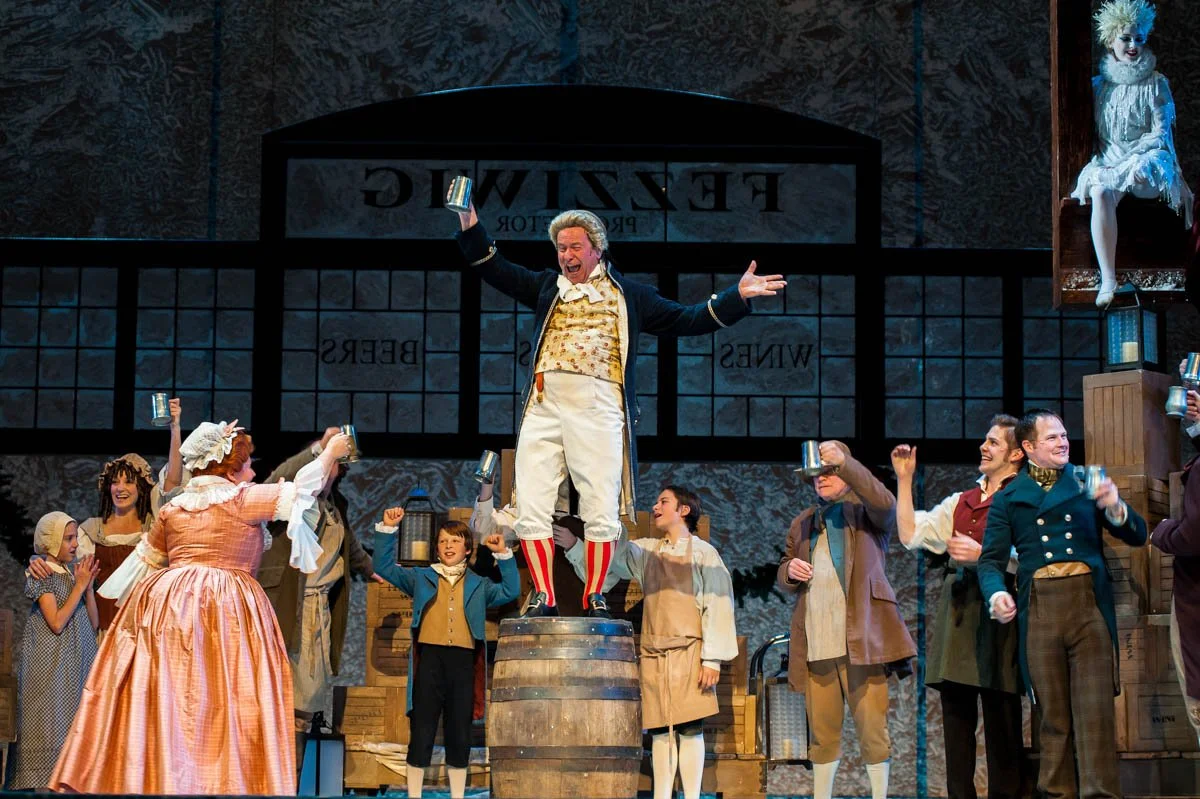 The cast of a theatrical performance celebrates on stage with some holding metal mugs. One man stands on a barrel with arms raised, wearing historical costume with striped socks. There are women and children dressed in period attire, and a figure wit