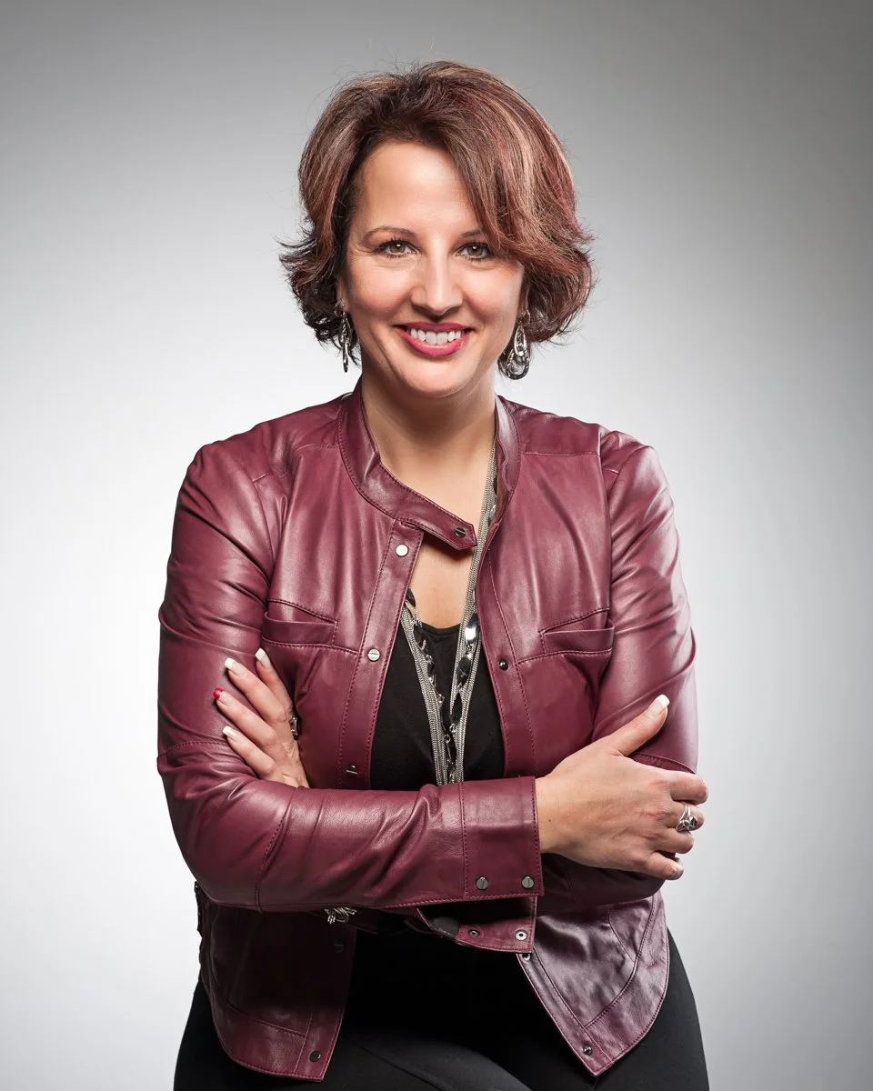 A woman with short, curly reddish-brown hair, smiling, wearing a maroon leather jacket over a black top, and posing with arms crossed against a plain gray background.