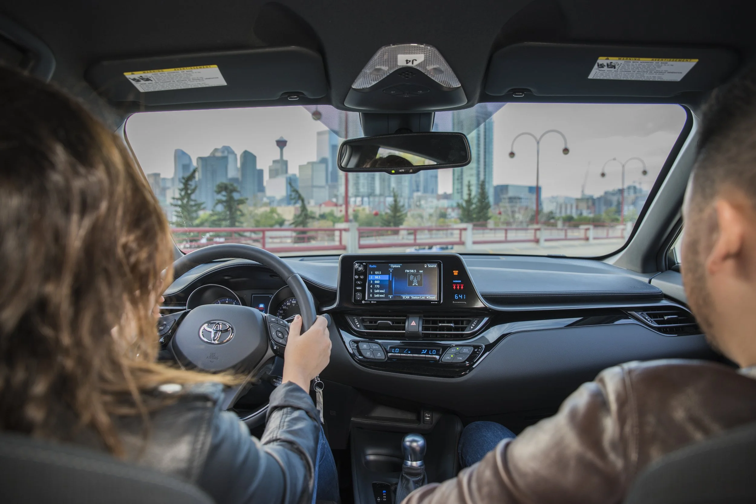 Interior of a car showing a woman driving and a man passenger, city skyline visible through the windshield.
