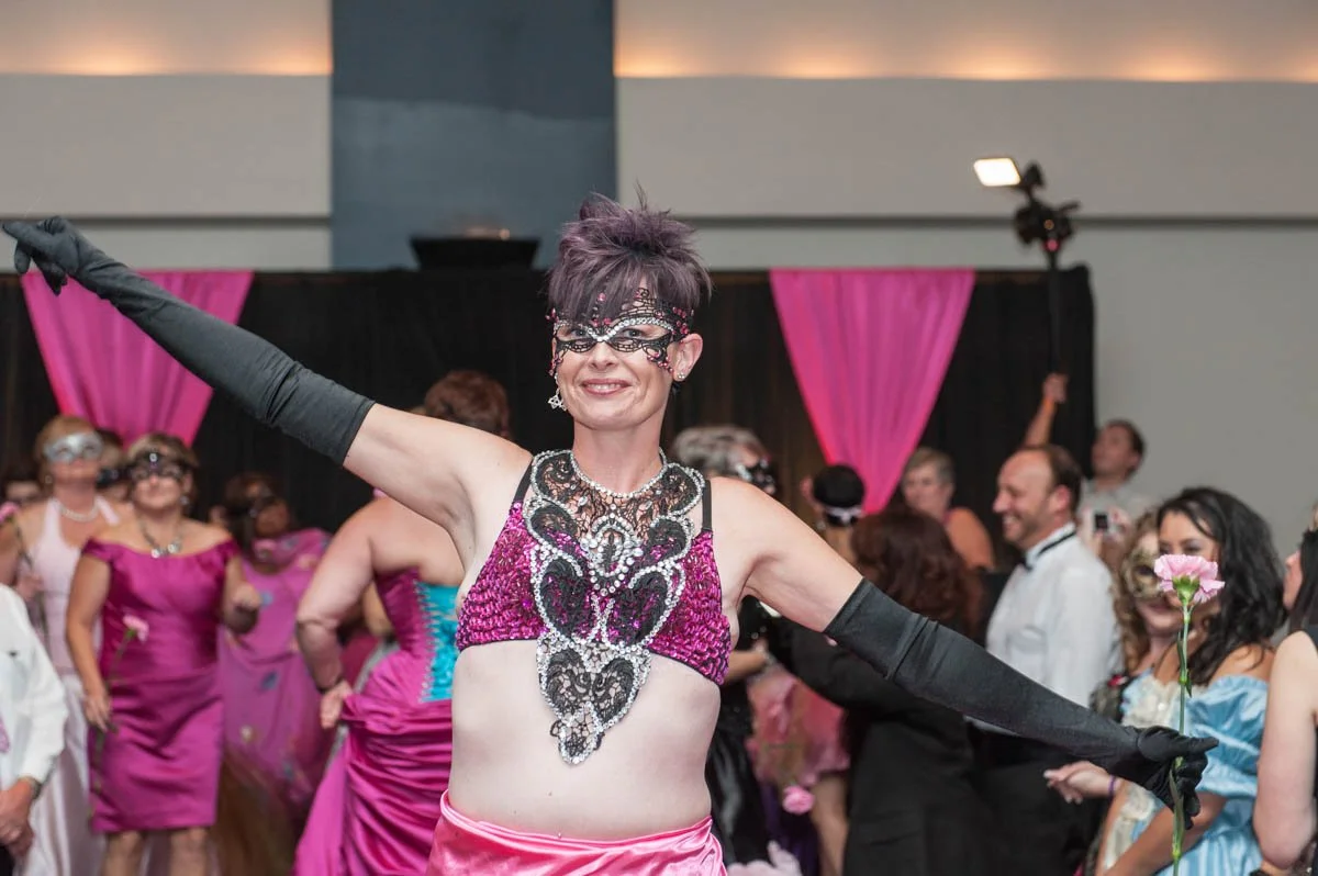 Woman in a colorful, sparkly costume and mask dancing at a party or celebration, surrounded by other people in formal and costume attire, with pink curtains and a lively atmosphere.
