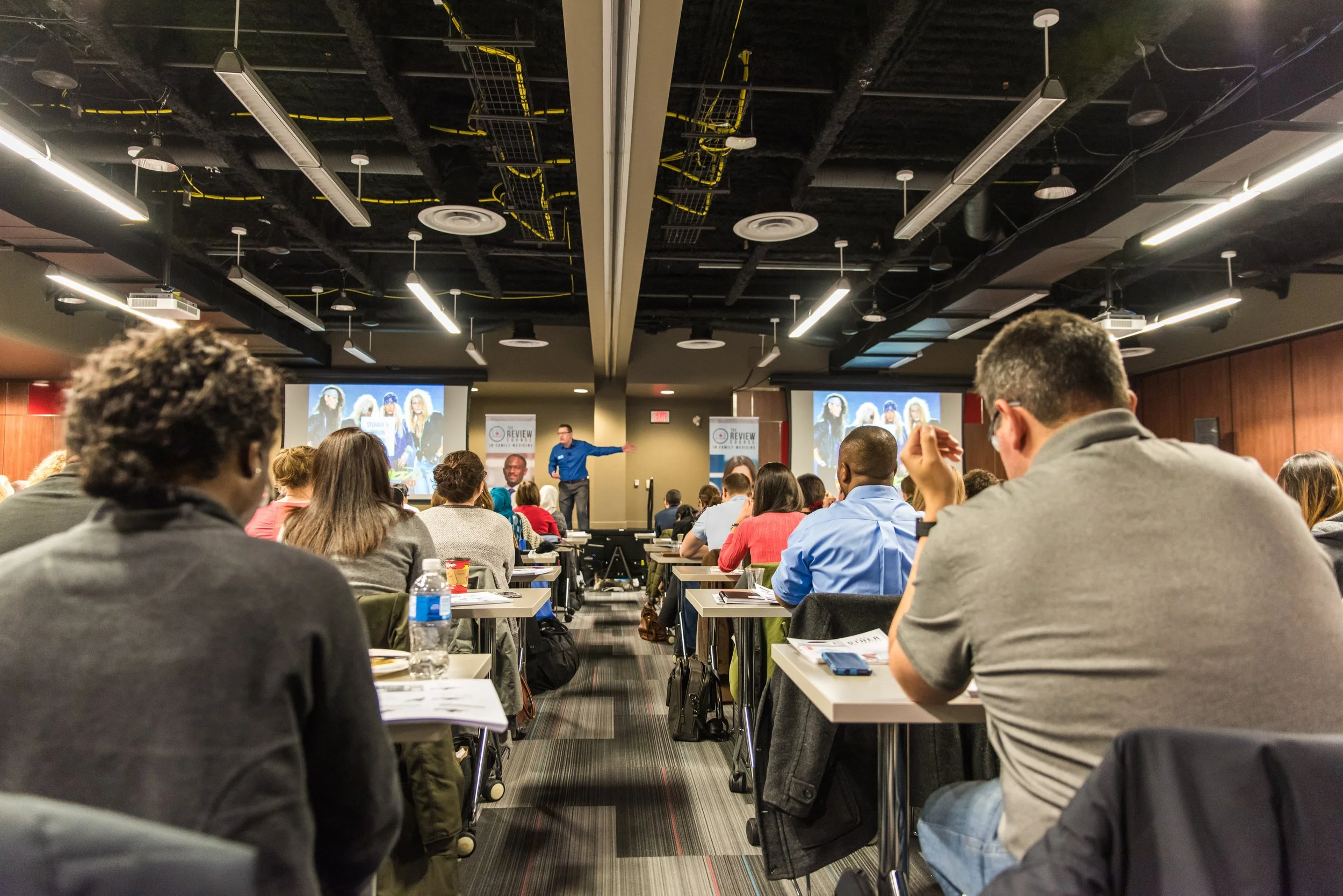Conference room filled with attendees taking notes while a speaker presents at the front, with two large screens displaying a group photo behind him.