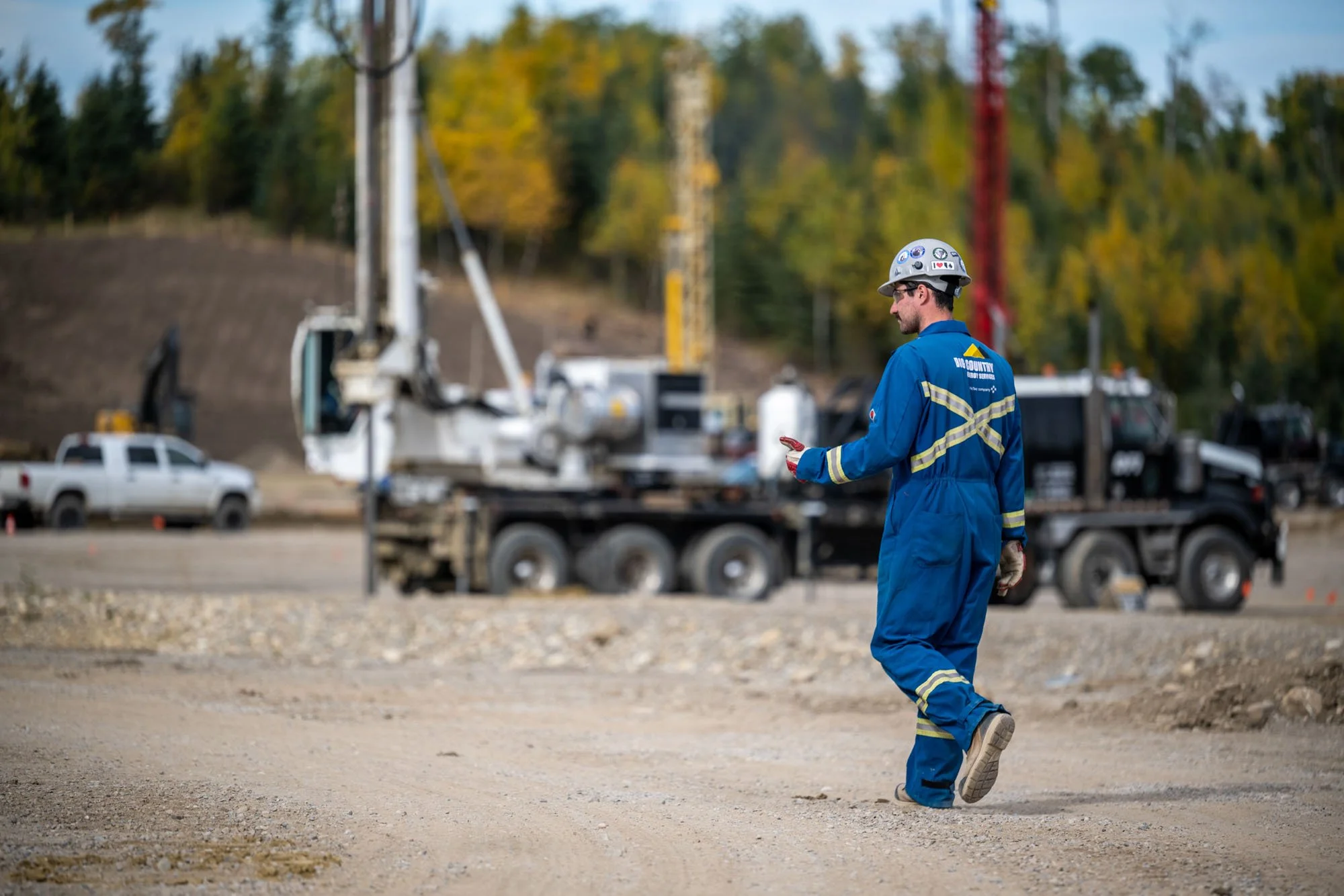 Worker in blue uniform and safety helmet walking on a construction or utility site with trucks and equipment in the background, trees with fall foliage visible.