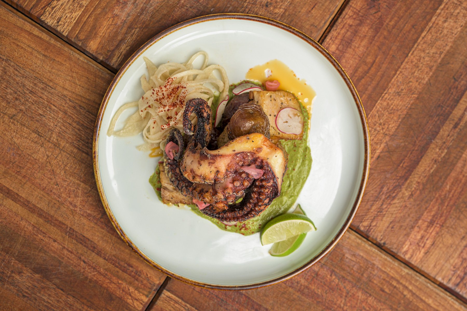 A plate of Mexican grilled octopus with noodles, sliced radishes, and lime wedges on a wooden table.