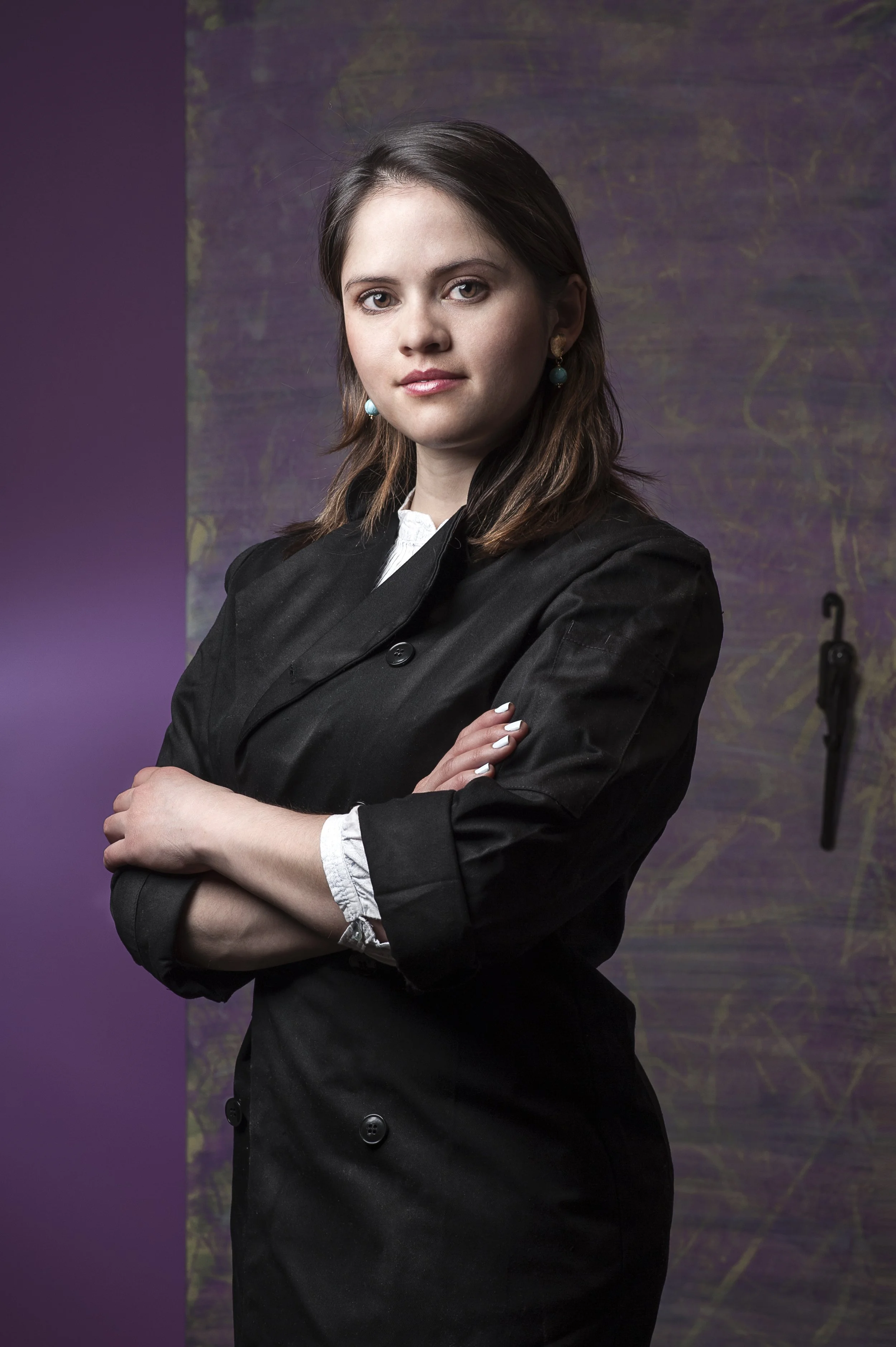 A woman with shoulder-length brown hair, wearing a black blazer with rolled-up sleeves, standing with her arms crossed and looking at the camera, with a purple and green abstract background.