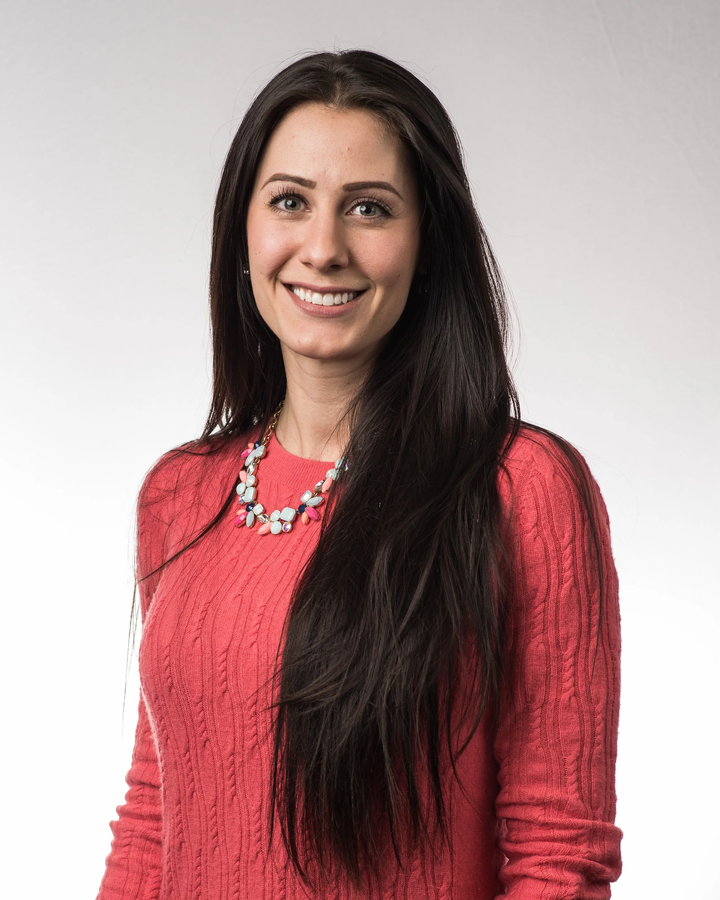 A smiling woman with long dark hair wearing a pink sweater and a colorful necklace, standing against a plain white background.