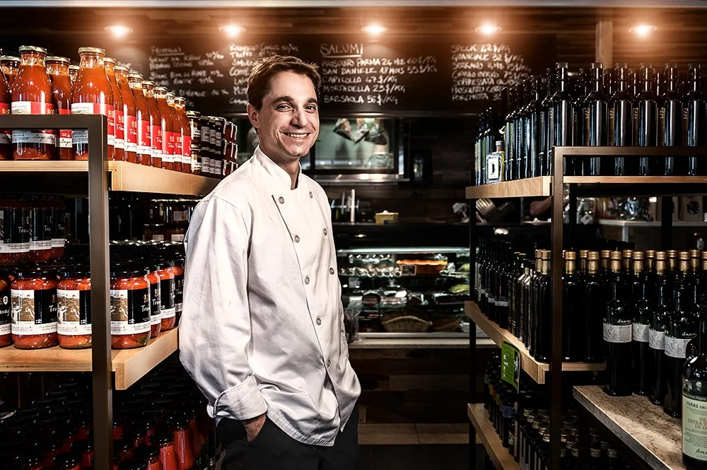 A smiling chef in a white coat stands in a grocery or specialty food store aisle surrounded by shelves of bottled tomato sauces and olive oils.