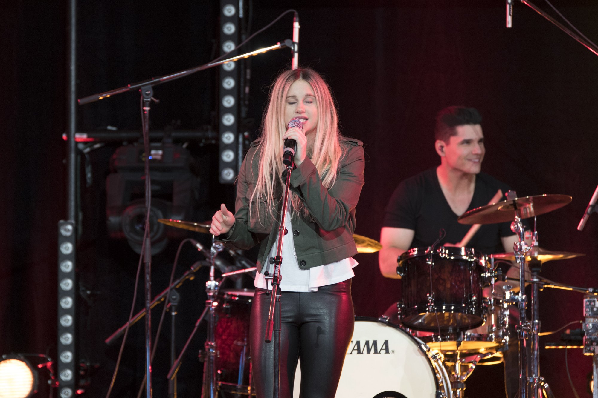 A woman singing into a microphone on stage with a drummer playing behind her. The woman has long blonde hair, is wearing a green jacket and black leather pants. The drummer is smiling and wearing a black shirt, with the drum set branded 'TAMA' visibl