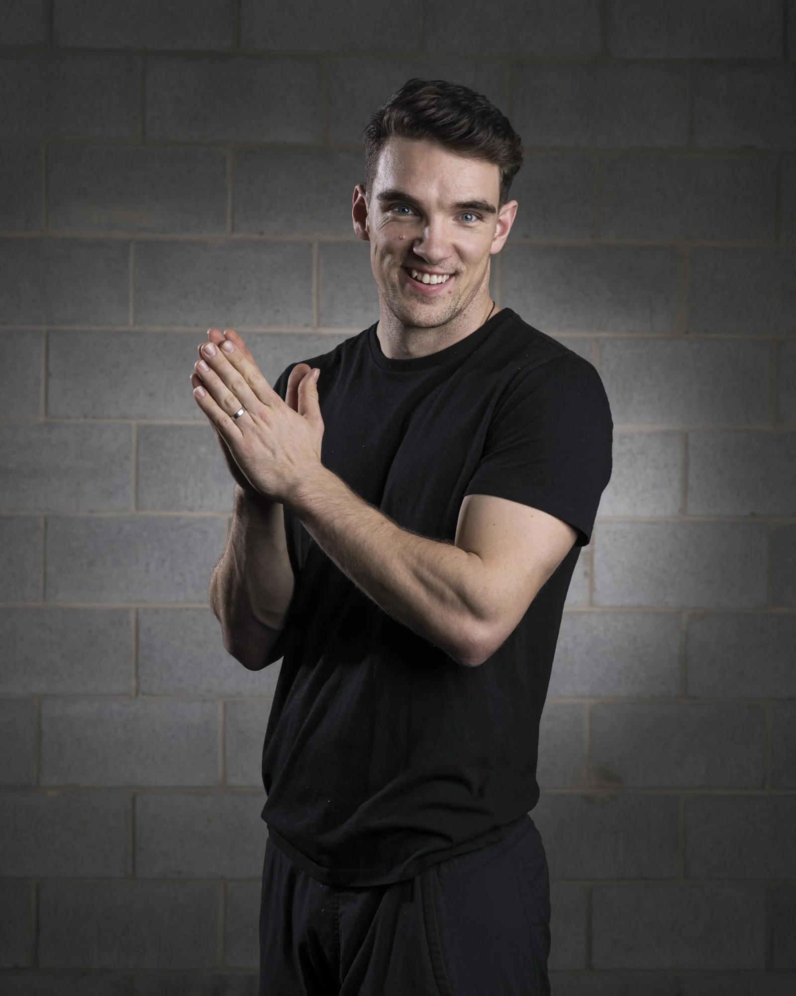 A smiling young man wearing a black t-shirt and black pants standing against a gray brick wall. He has his hands together in a prayer-like pose.