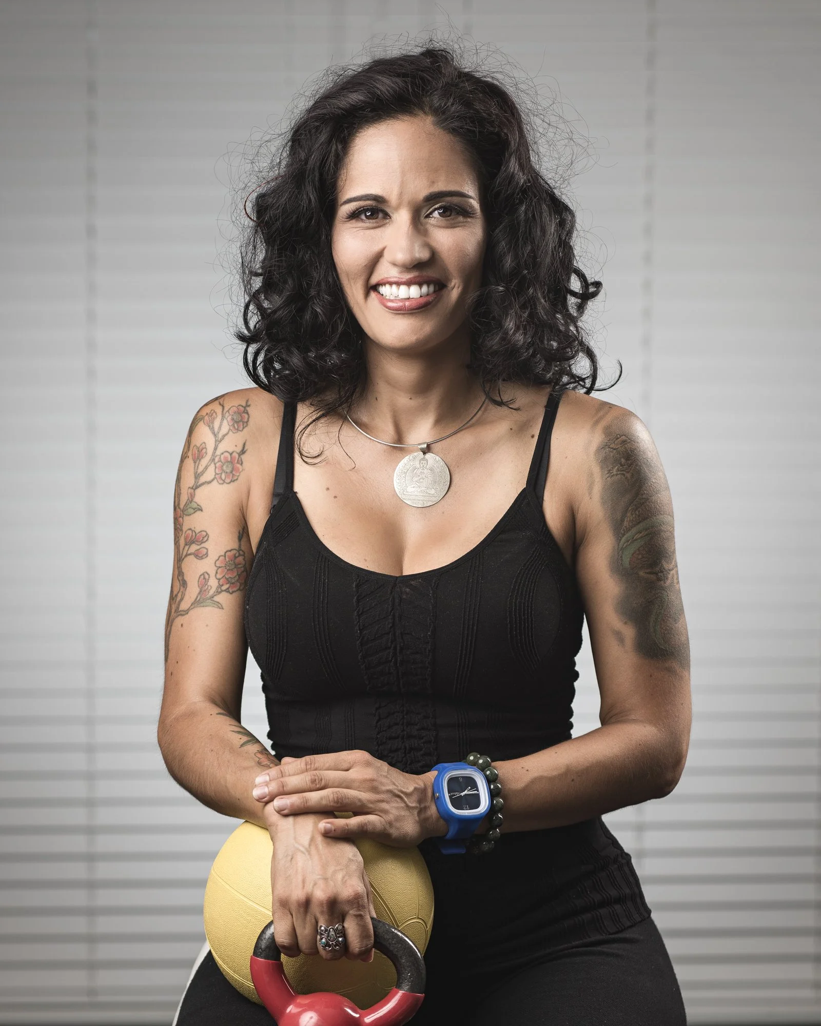 A smiling woman with tattoos on her arms, wearing a black tank top, a silver necklace, and a blue fitness watch, sitting with her hands on a kettlebell and medicine ball, against a neutral background.