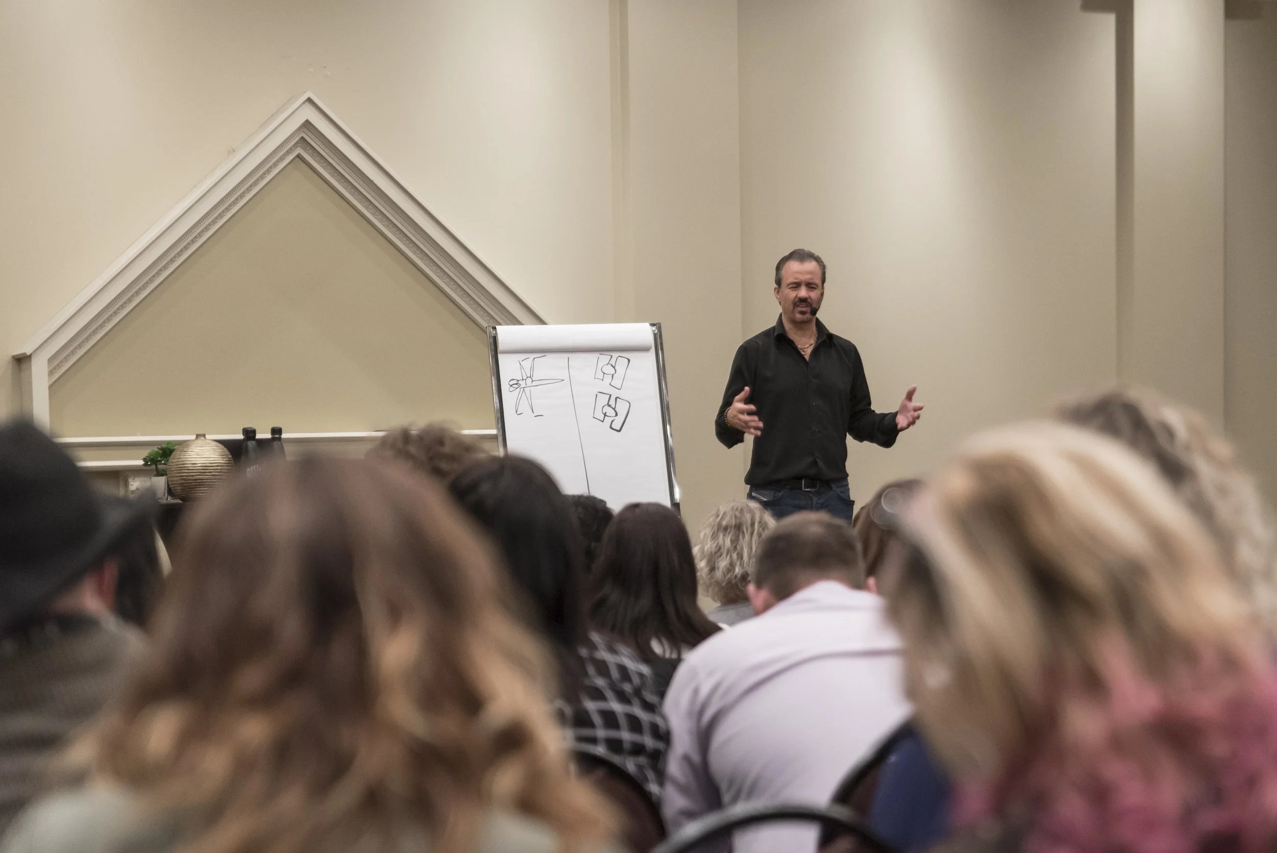 A man is giving a presentation to an audience in a conference room, standing in front of a whiteboard with sketches on it.