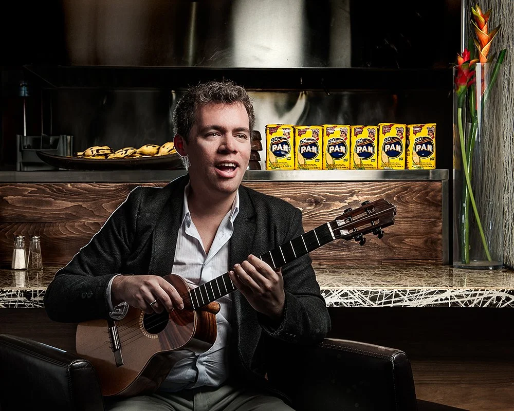 Man playing guitar and singing in a cafe or restaurant with a countertop behind him, featuring bananas, juice boxes, salt and pepper shakers, and a vase with flowers.