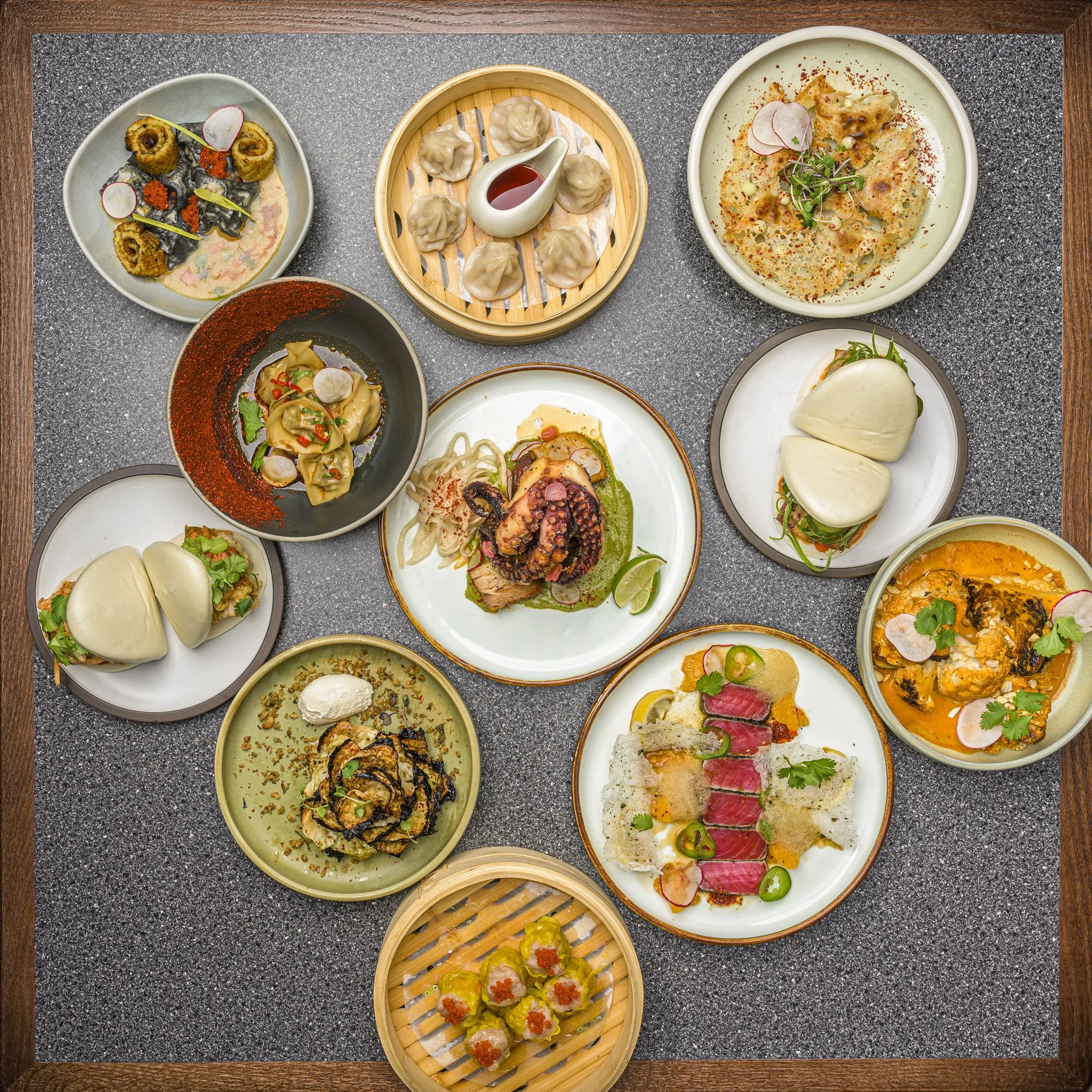 A top-down view of a variety of Asian dishes, including dumplings, sushi, and other small plates, served on a dark, textured tabletop.