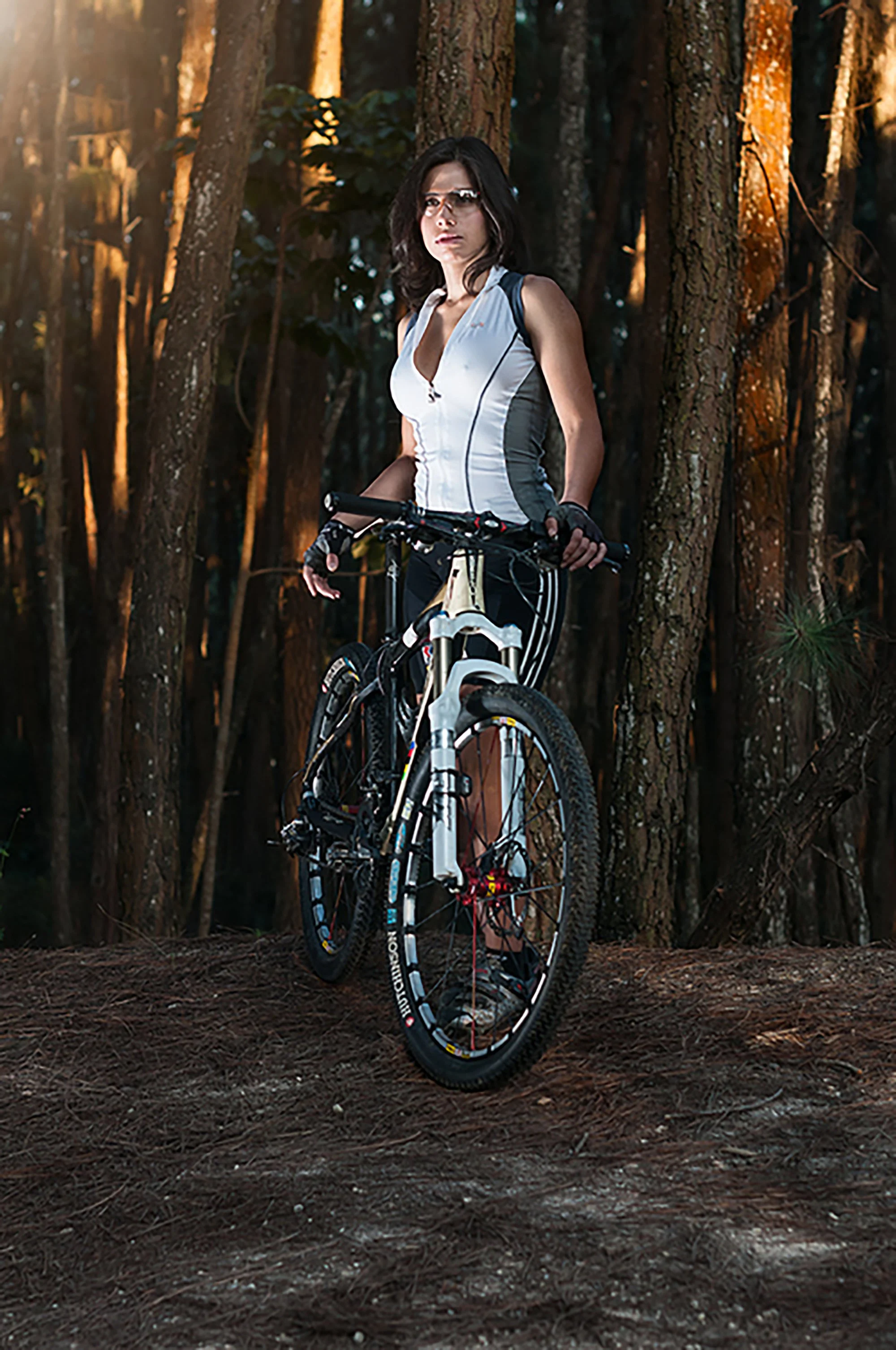 A woman in athletic gear stands with a mountain bike in a wooded forest at sunset.