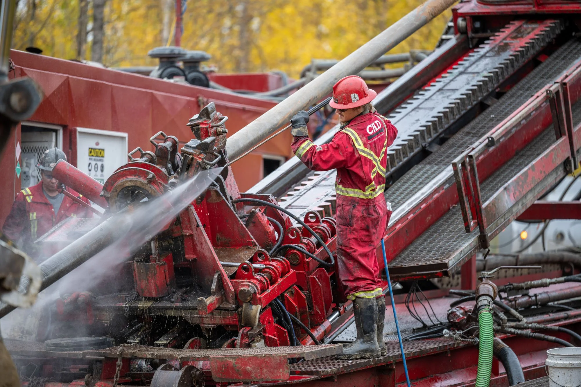 Firefighter in red uniform and helmet working on large red firefighting equipment outdoors with trees in the background.