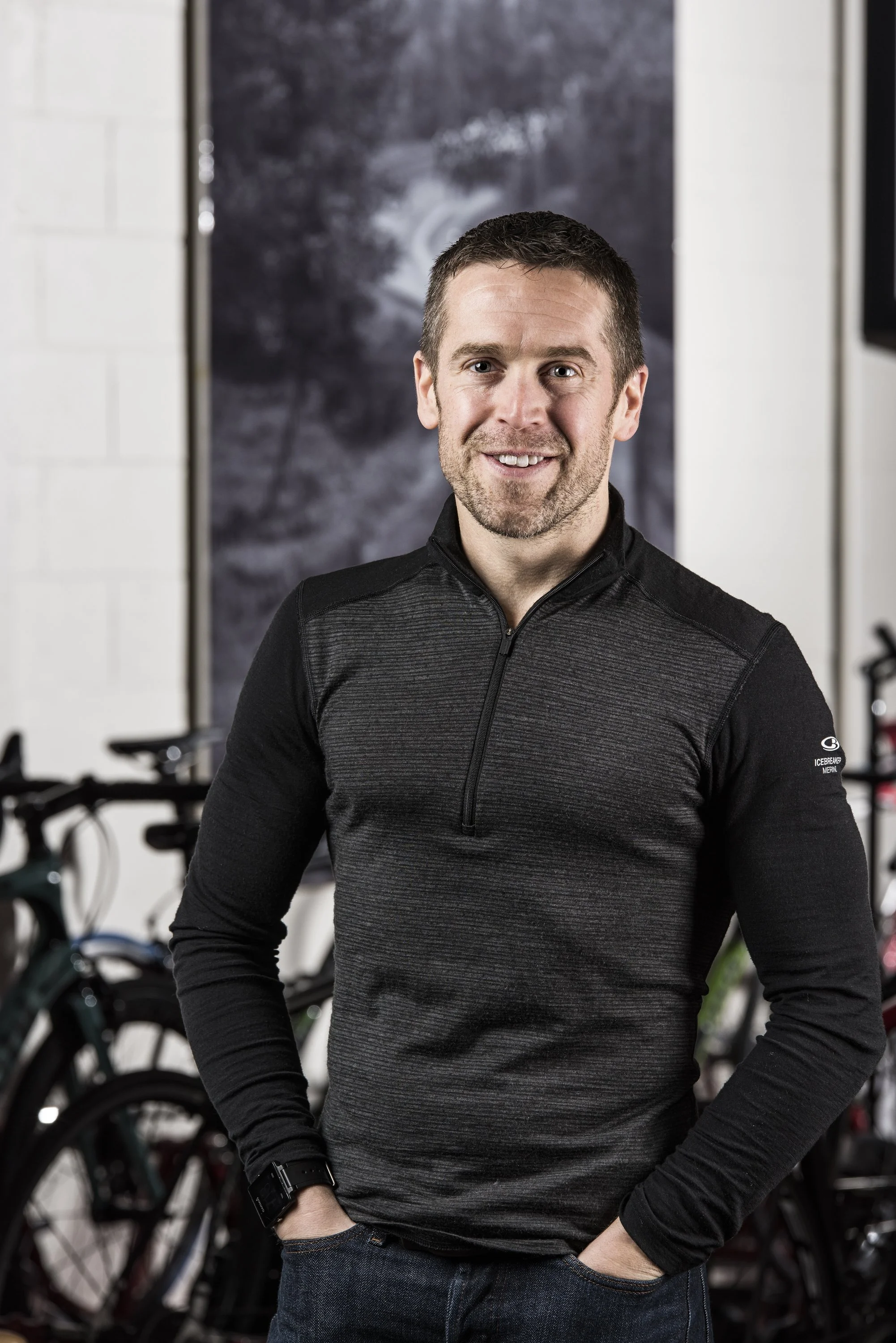 A man wearing a black athletic pullover with a quarter-zip, standing in front of bicycles in a gym or bike shop, smiling at the camera.