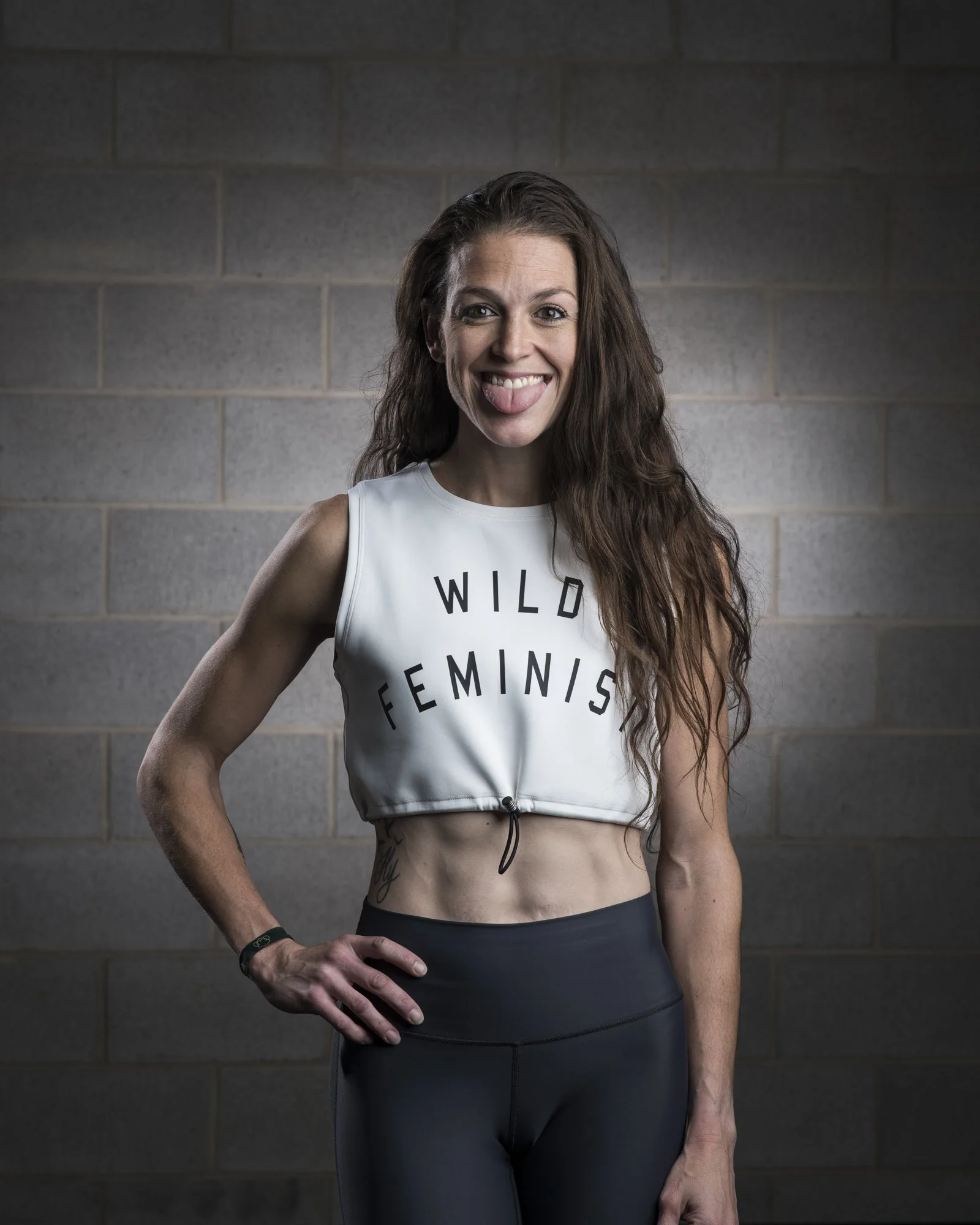 A woman with long, wavy brown hair and a big smile sticking her tongue out, wearing a white crop top with the text 'WILD FEMINIST' printed on it and black leggings, standing against a gray brick wall.